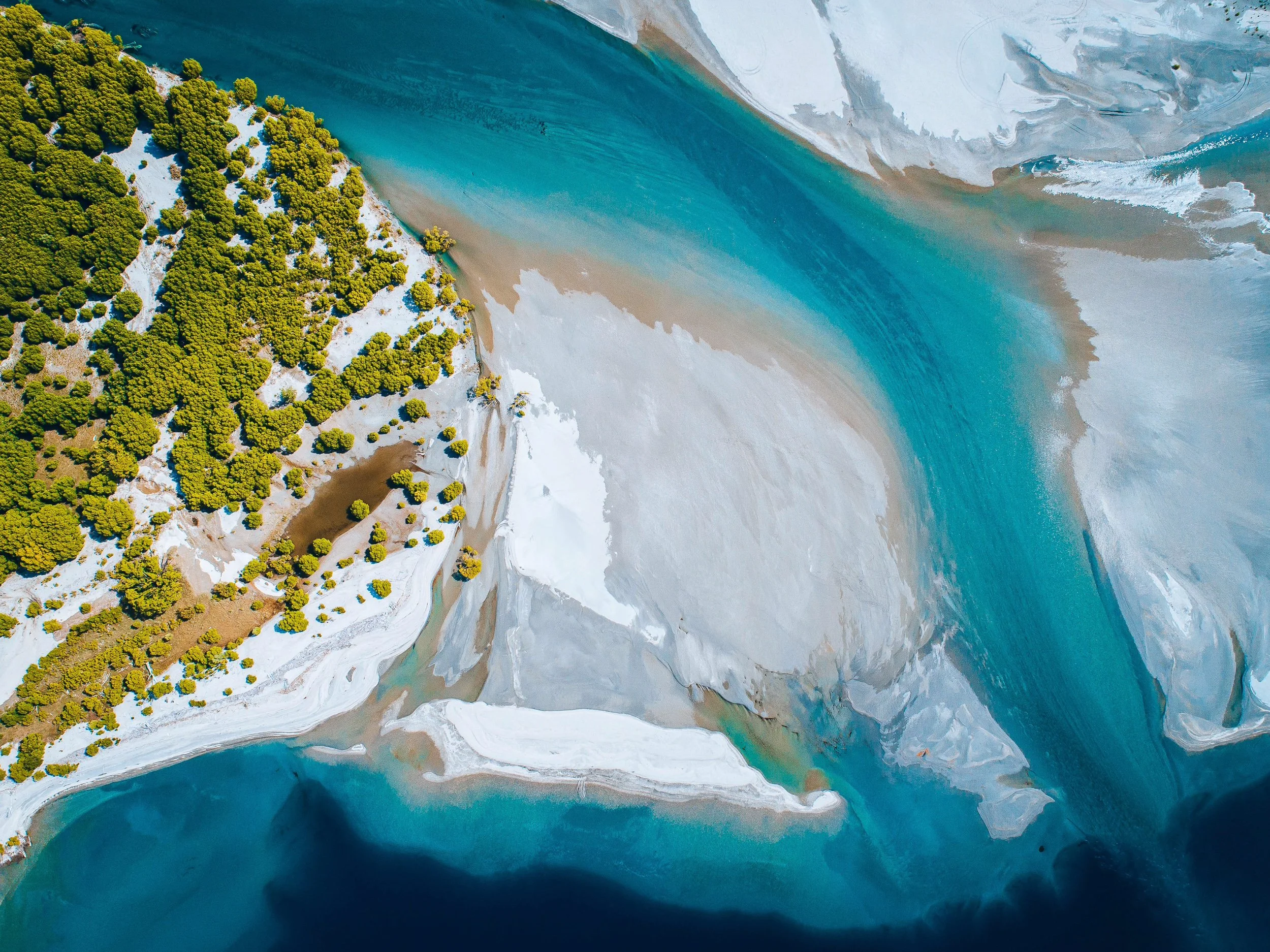Aerial view of a river with turquoise water flowing past a snowy, white, and rocky landscape. Green trees are on one side of the landscape.