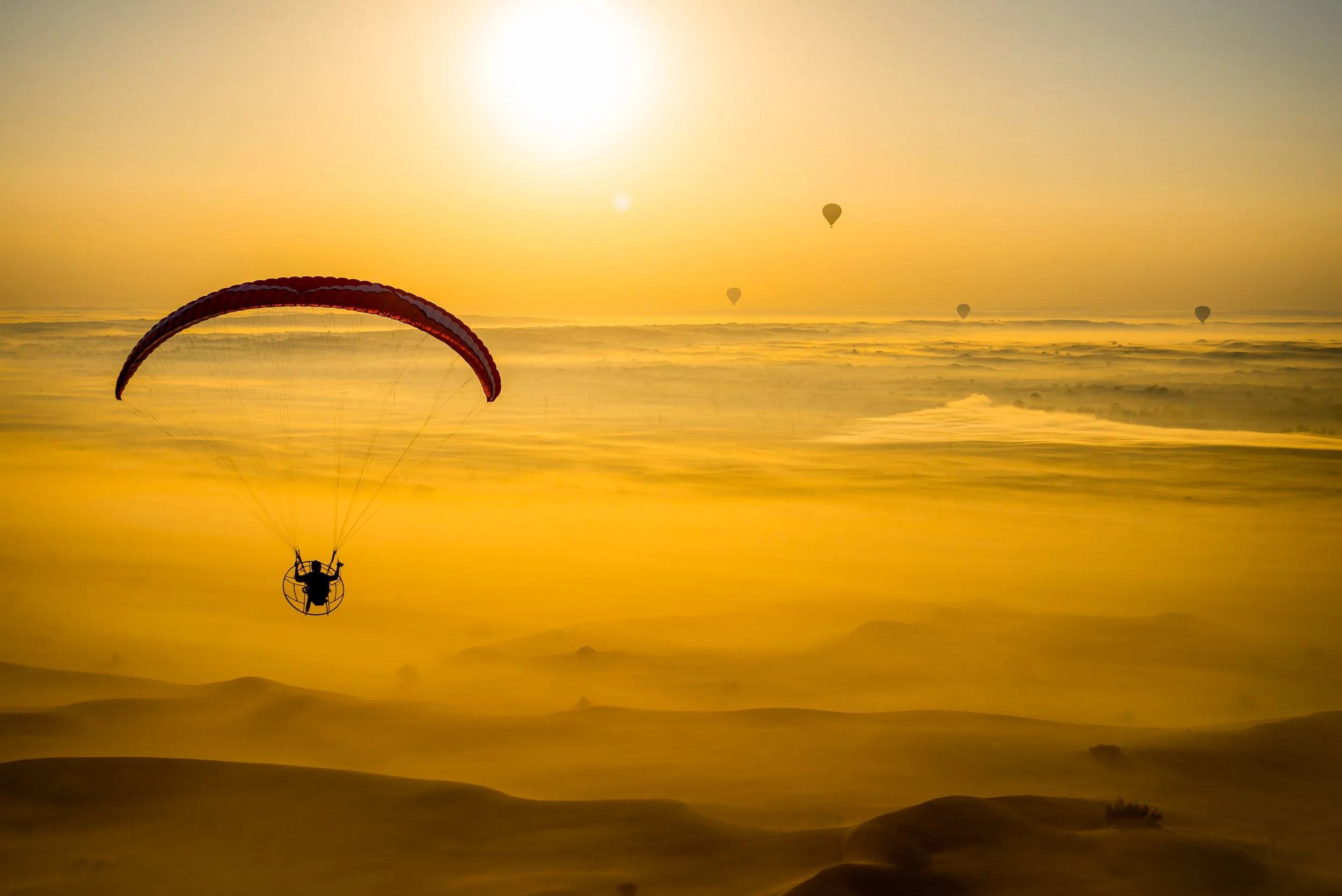 A person paragliding over sand dunes during a golden sunset, with several hot air balloons in the sky.