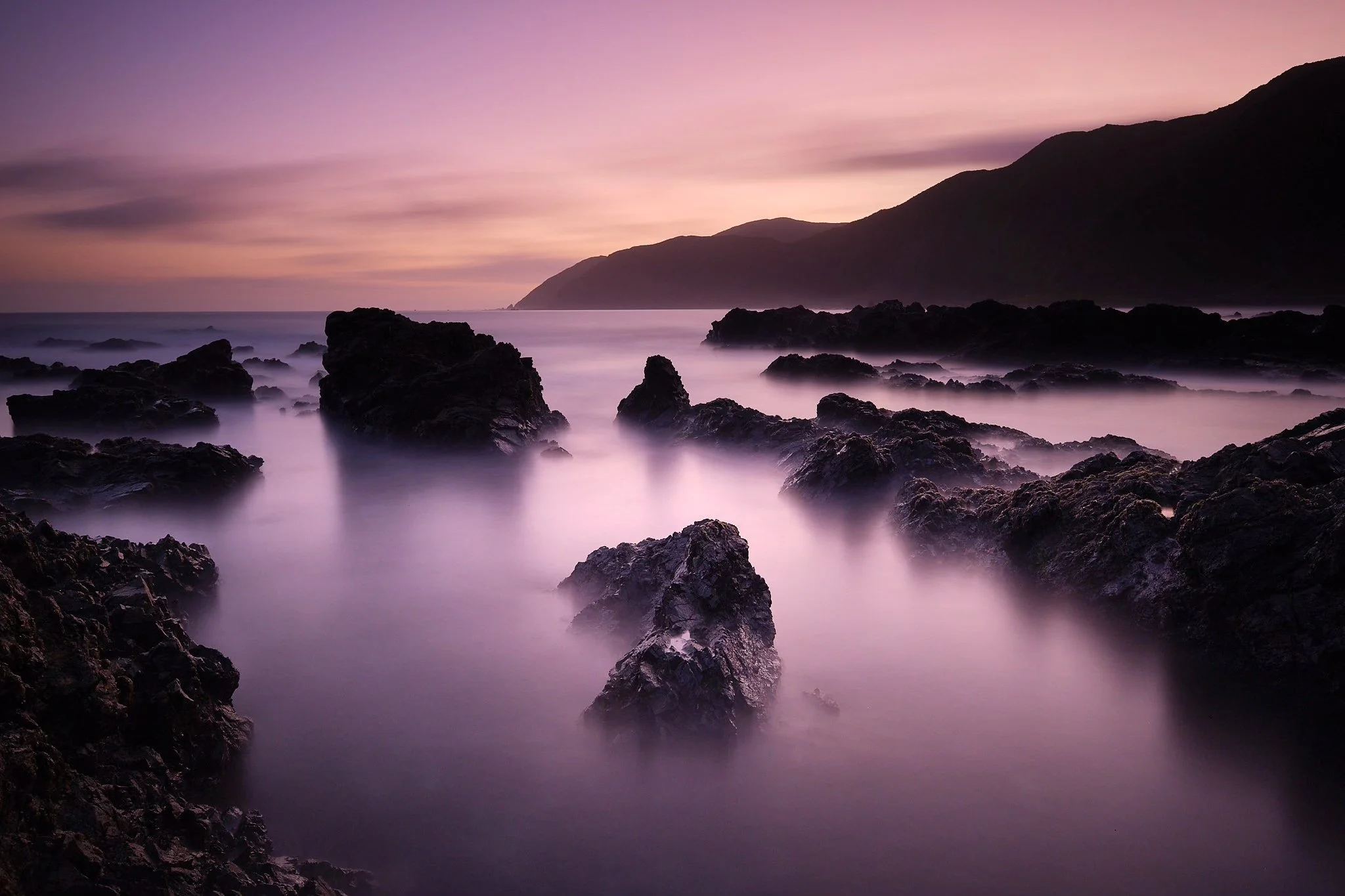 A long exposure photo of a rocky coastline at sunset, with purple and pink sky and calm sea waves surrounding the rocks.