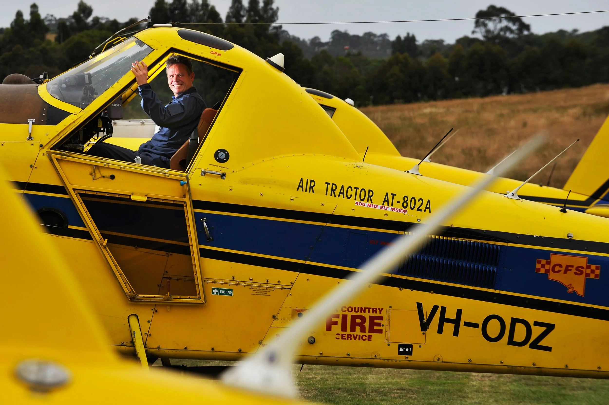 A man smiling and waving from the cockpit of a yellow and blue helicopter on a grassy field, with trees and hills in the background.