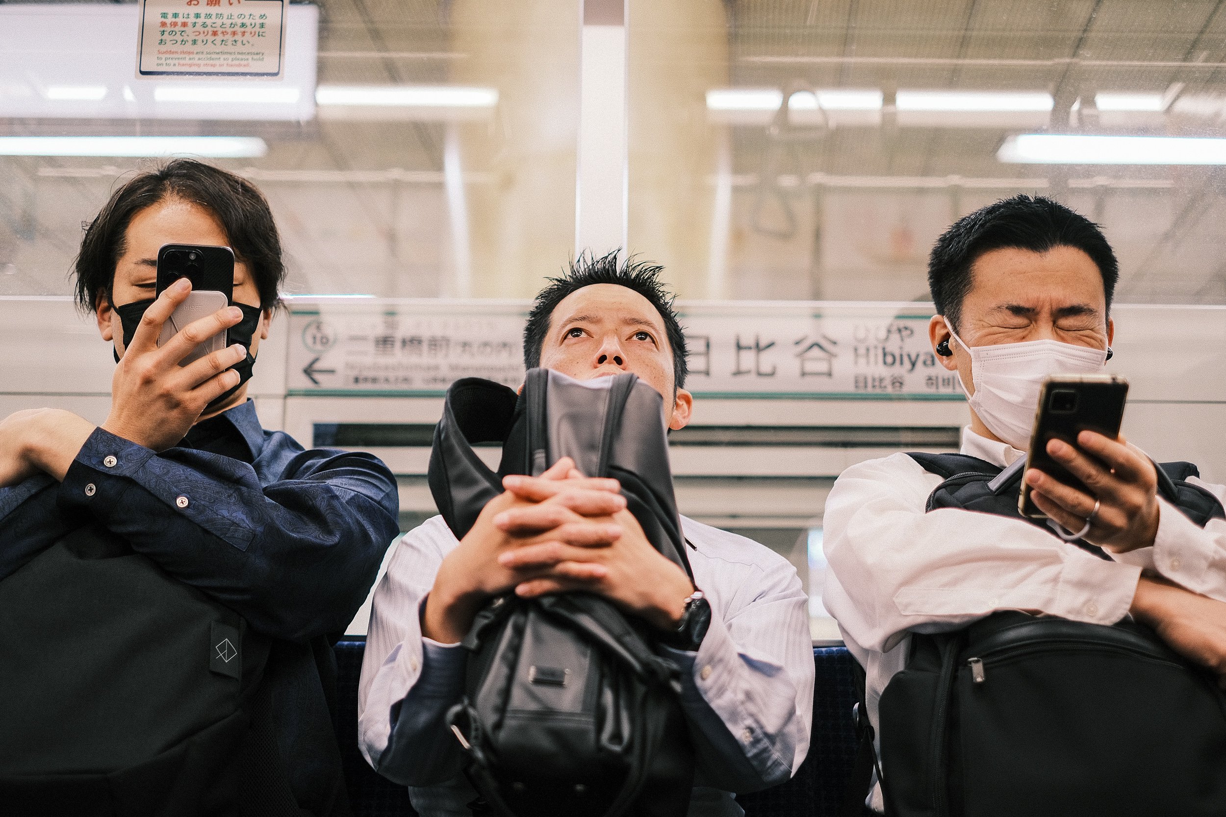 Three people wearing masks sitting on a train platform, with two using phones and one holding a backpack with hands clasped together.