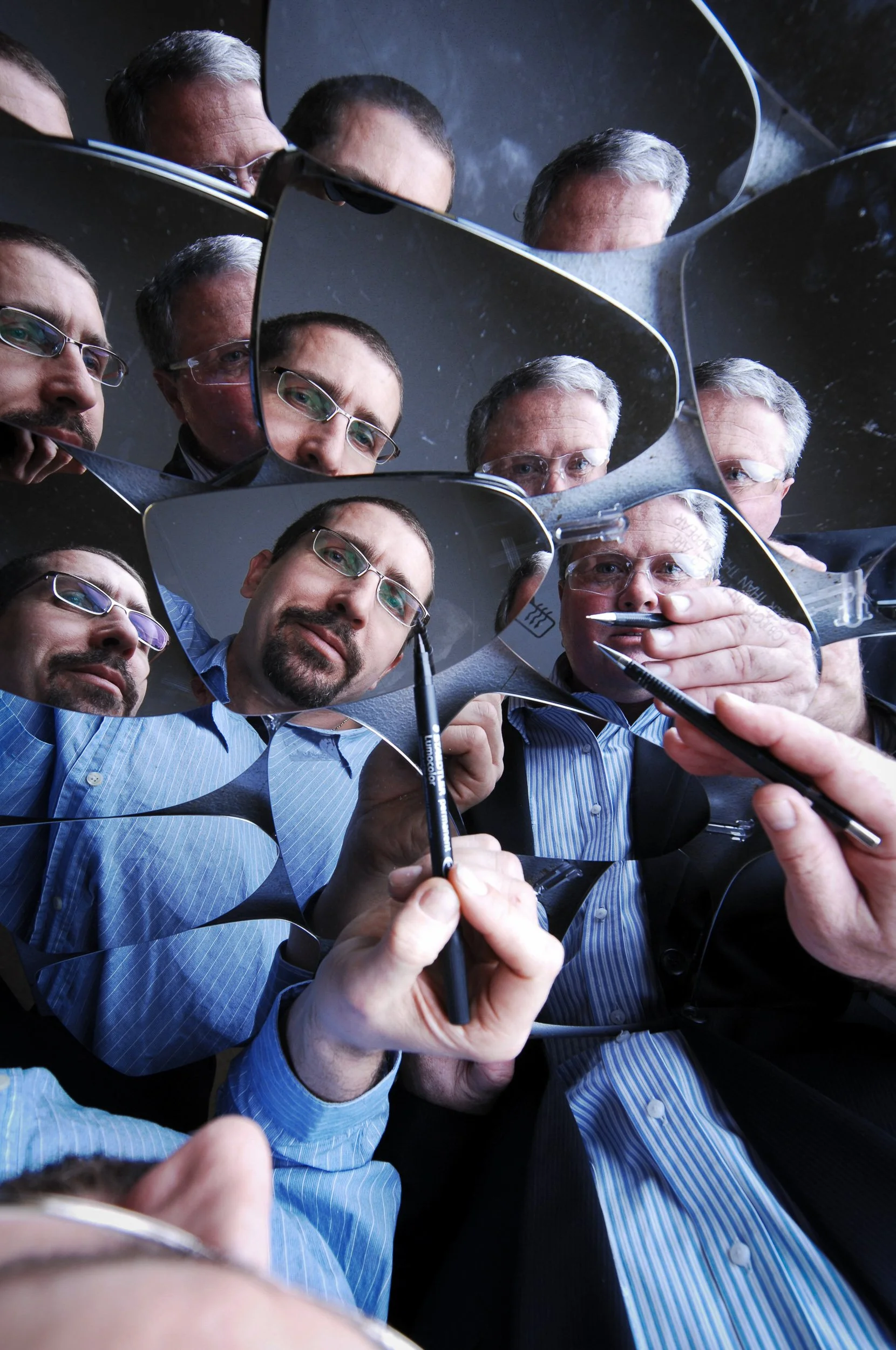 A group of men in business attire and glasses looking at broken mirror pieces, with reflections of their faces, as one man writes or draws with a black pen.