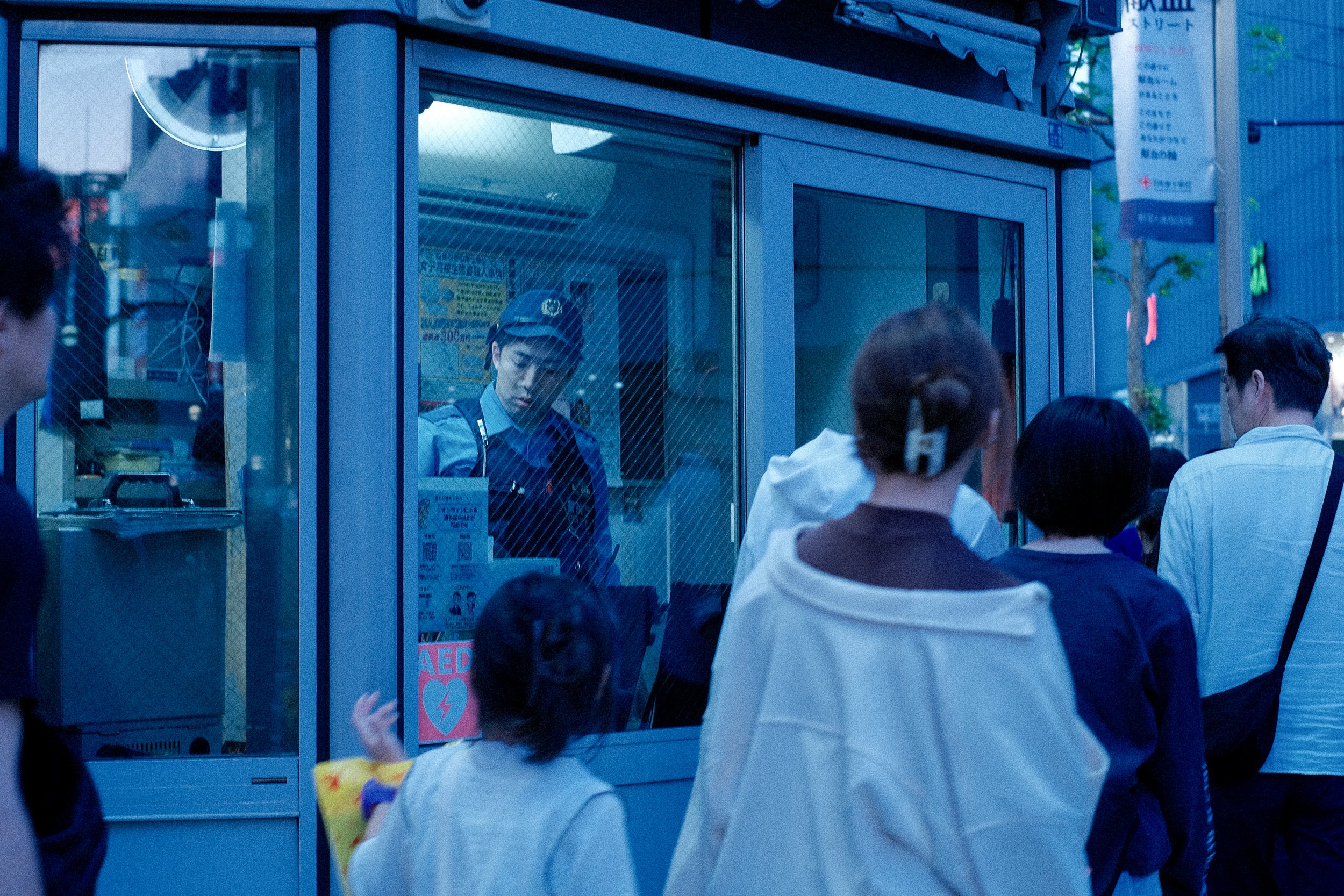 A group of people standing in line outside a small building, with a uniformed female worker inside the building viewed through a glass window.