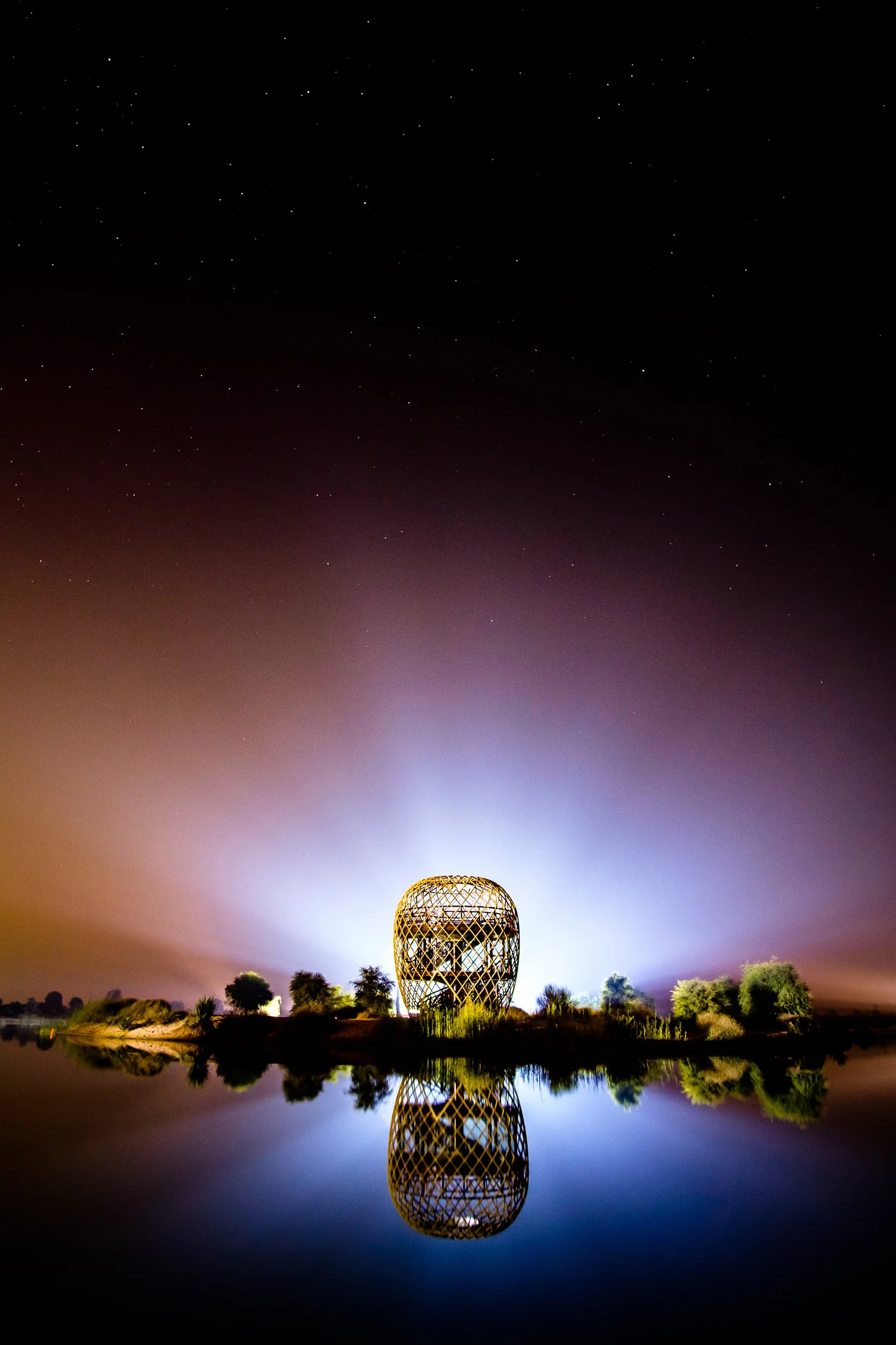 A glowing, open lattice structure shaped like a vase or helmet is located on a small island or peninsula, with its reflection visible in the water below. The sky is dark with visible stars, and there is a bright light behind the structure creating a 