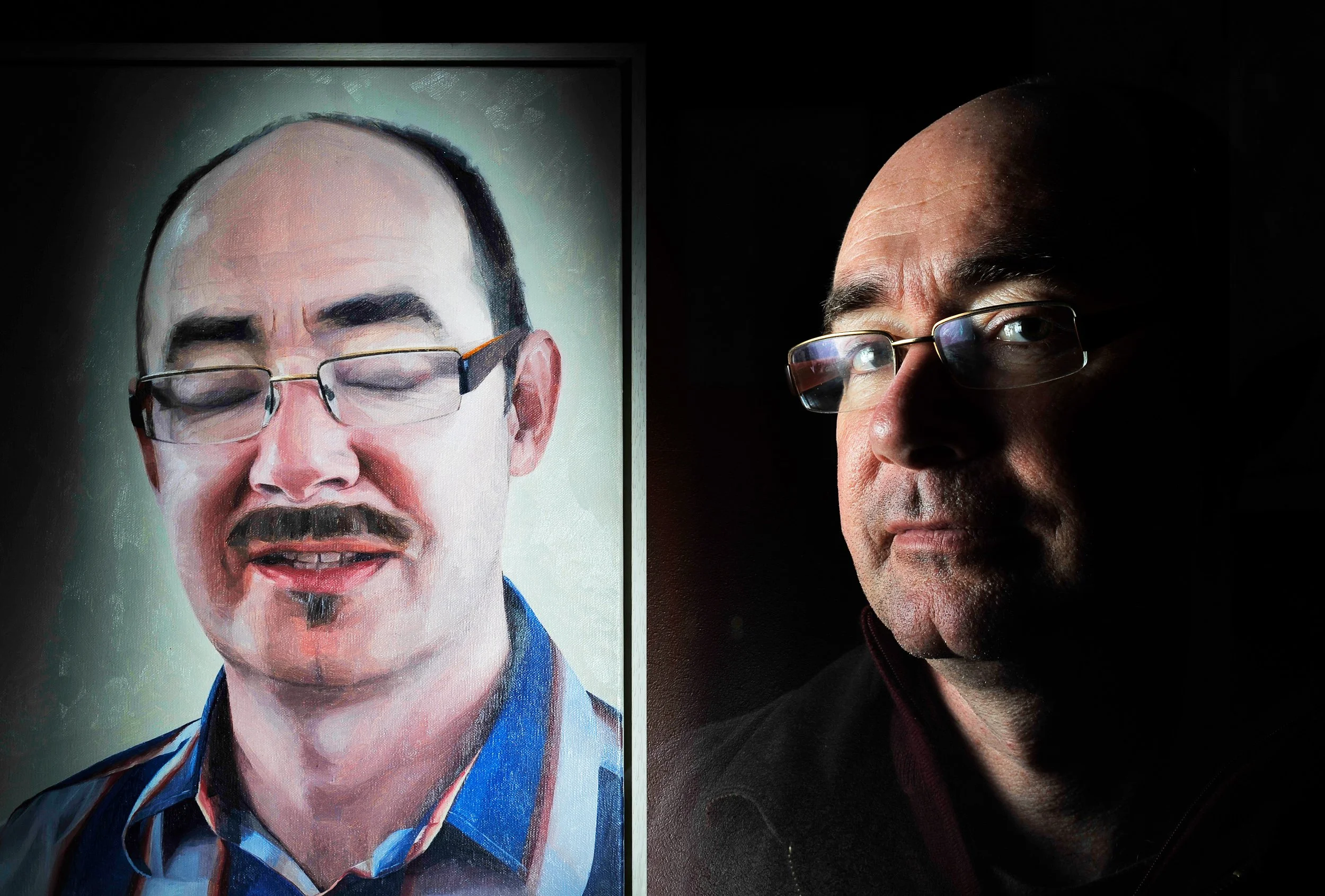 A portrait of a man with glasses in a dark setting, with a painting or image of a similar man with glasses and a mustache and goatee on the left.