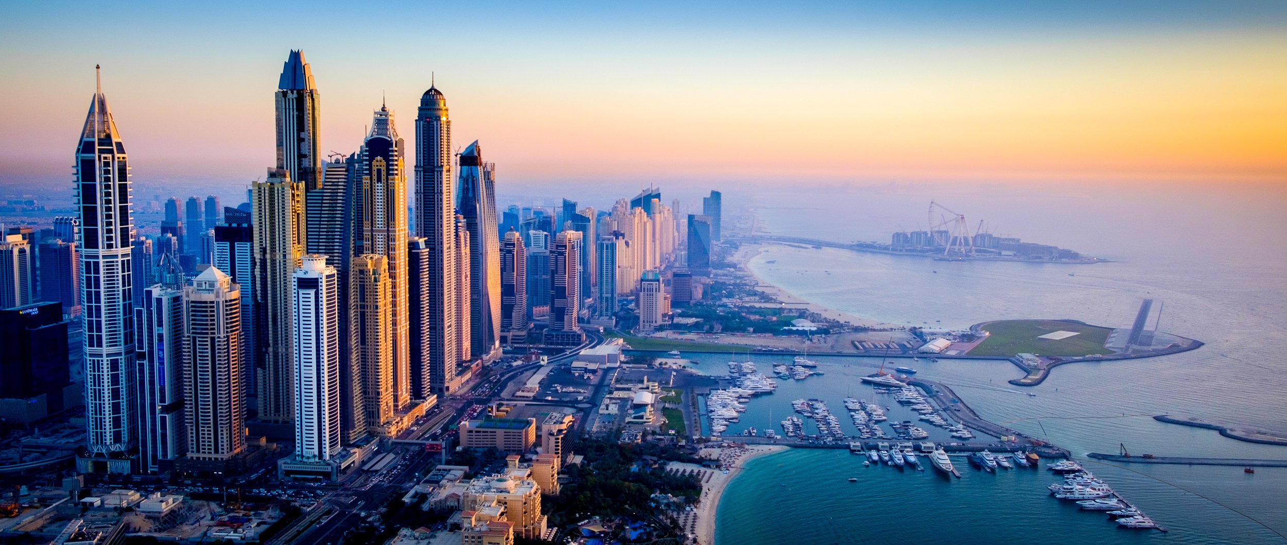Aerial view of a modern city skyline with tall skyscrapers near a marina, harbor, and beach at sunset.