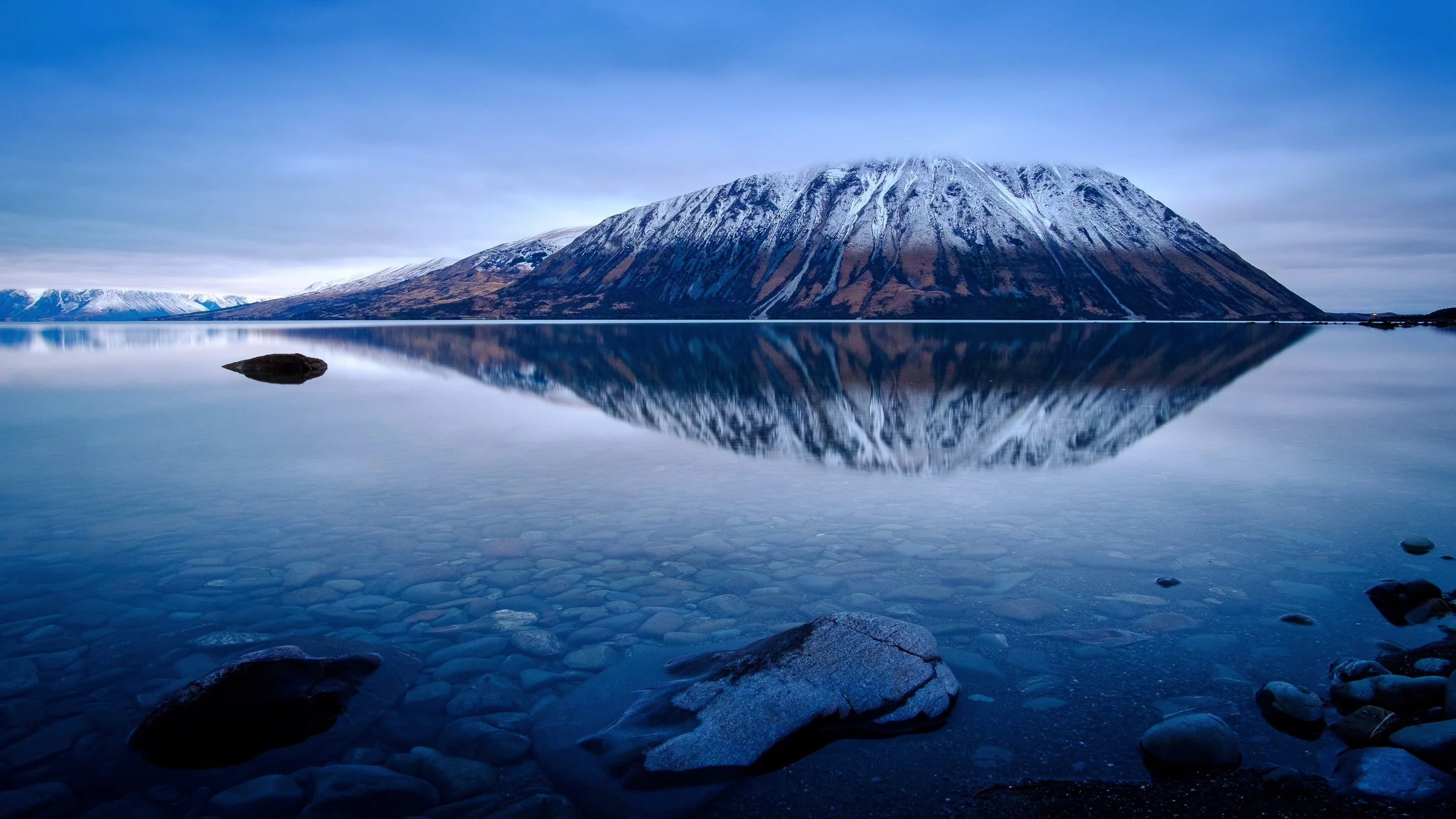 Snow-capped mountain reflected in calm lake with rocks along the shoreline.