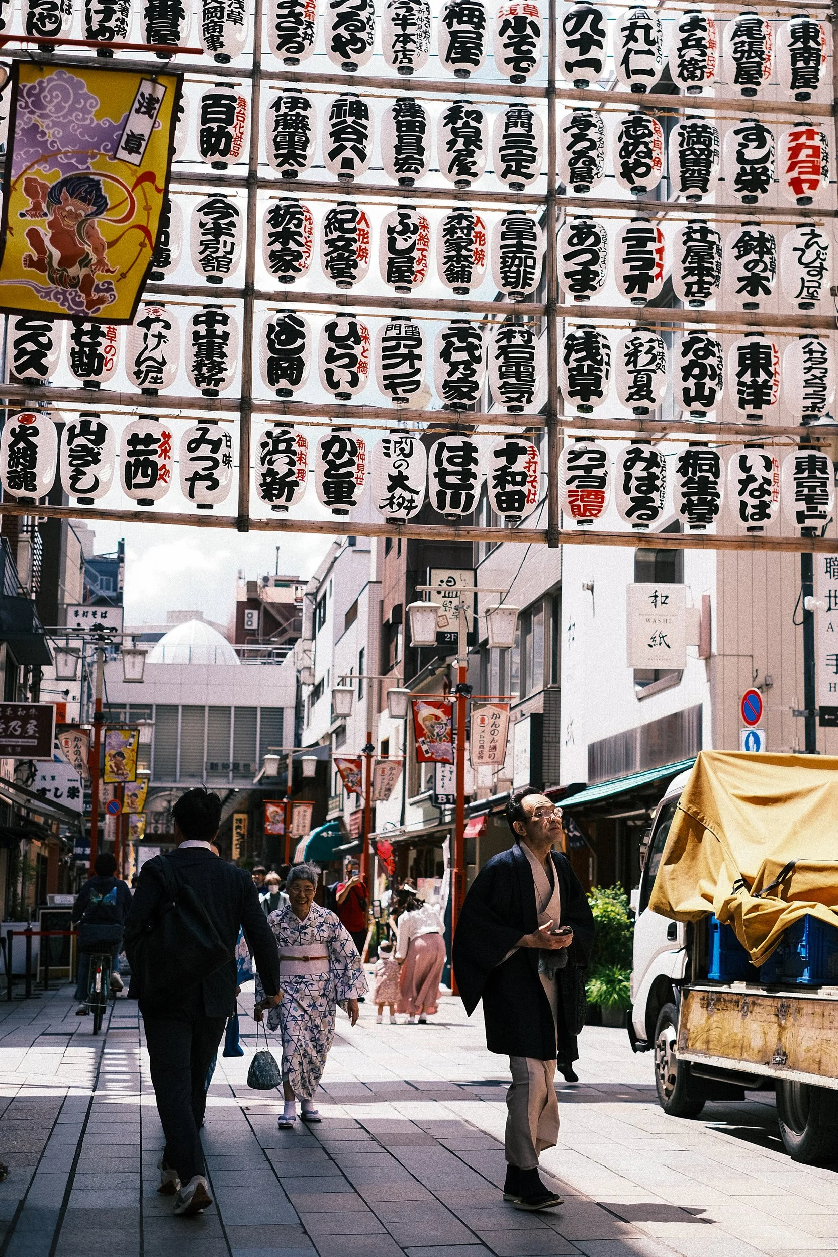 A busy street in Japan decorated with hanging paper lanterns and banners. People are walking, some in traditional kimonos and others in modern attire. A truck with a yellow cover is parked on the side.