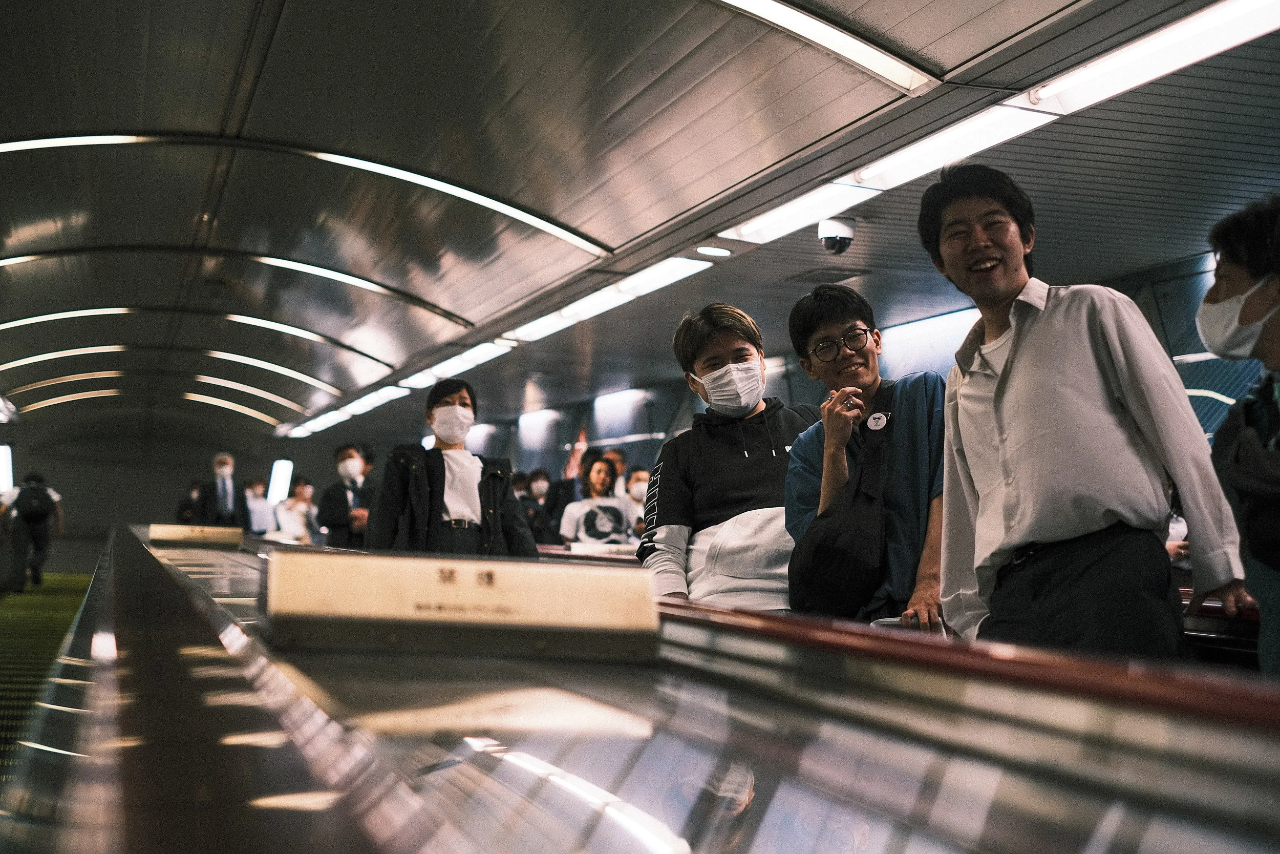 People wearing masks waiting in line at an underground transit station, with some smiling and engaging with each other.