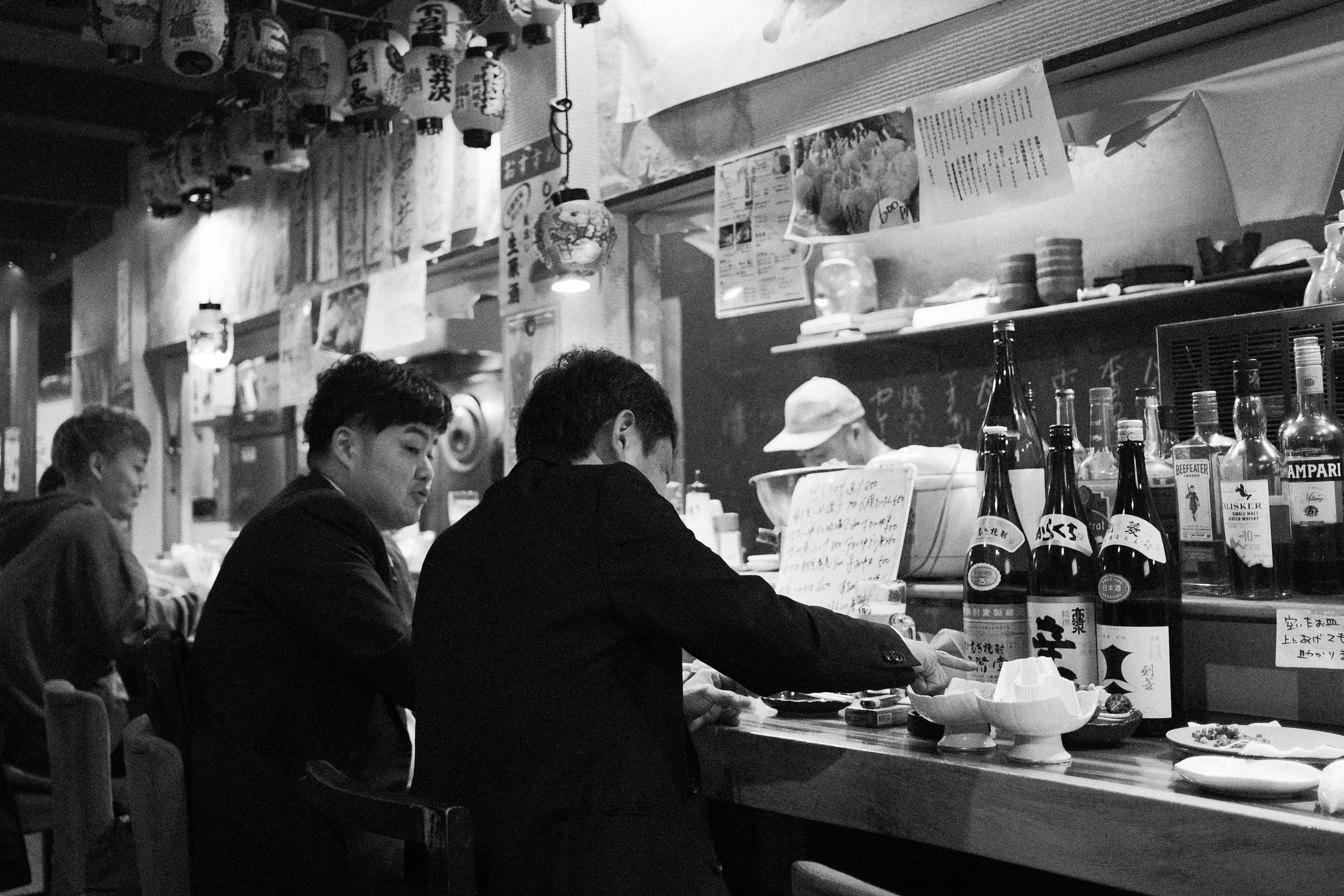 A black and white photo of a Japanese restaurant or bar with customers sitting at the counter. Three men are visible, two in suits and one with a cap, sitting in front of bottles and bowls, with a staff member behind the counter preparing food. The b