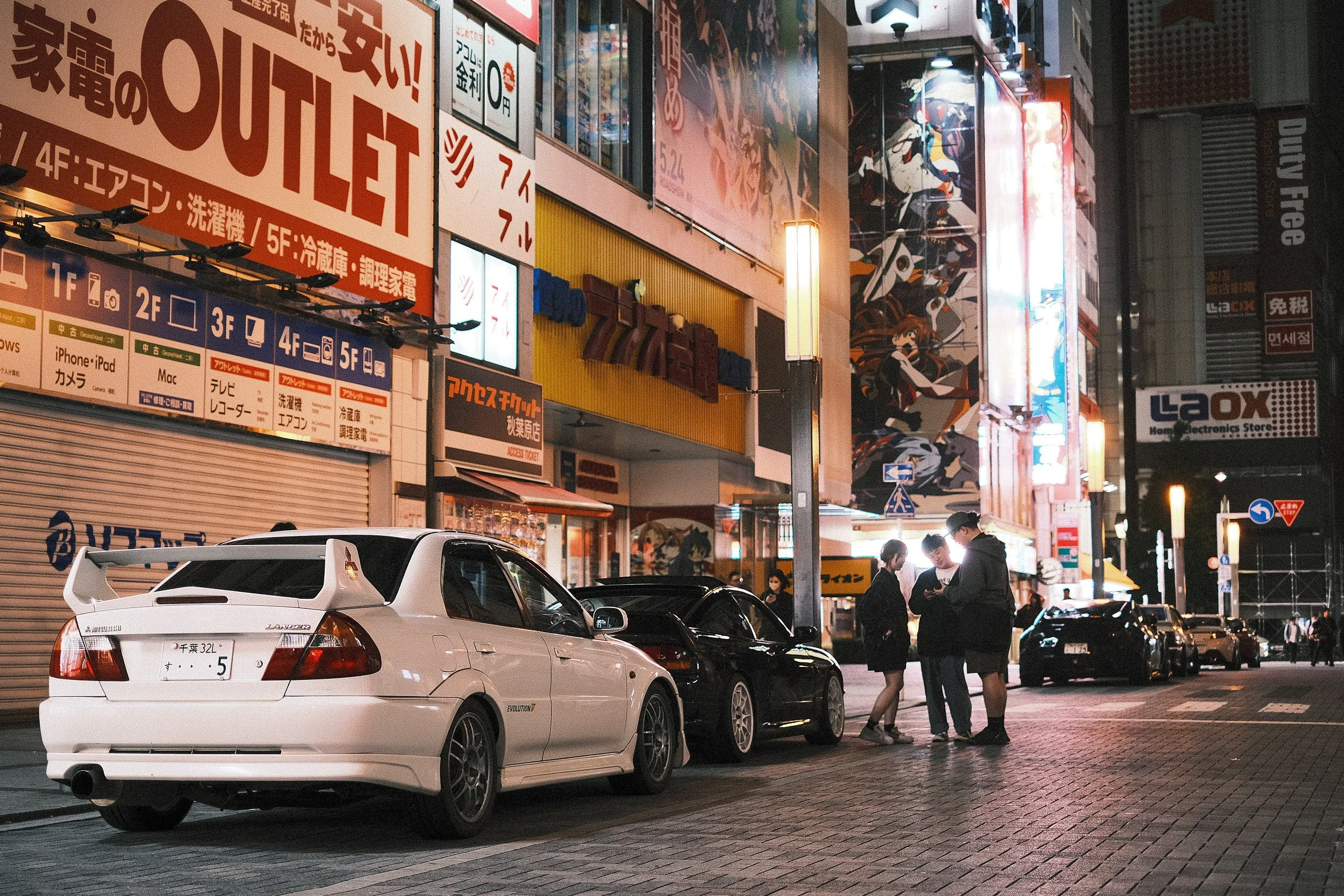 Nighttime street scene with parked cars and a group of four people standing and talking in front of brightly lit commercial buildings with large colorful signs and advertisements in Japanese, and comic or anime-style murals on the buildings.