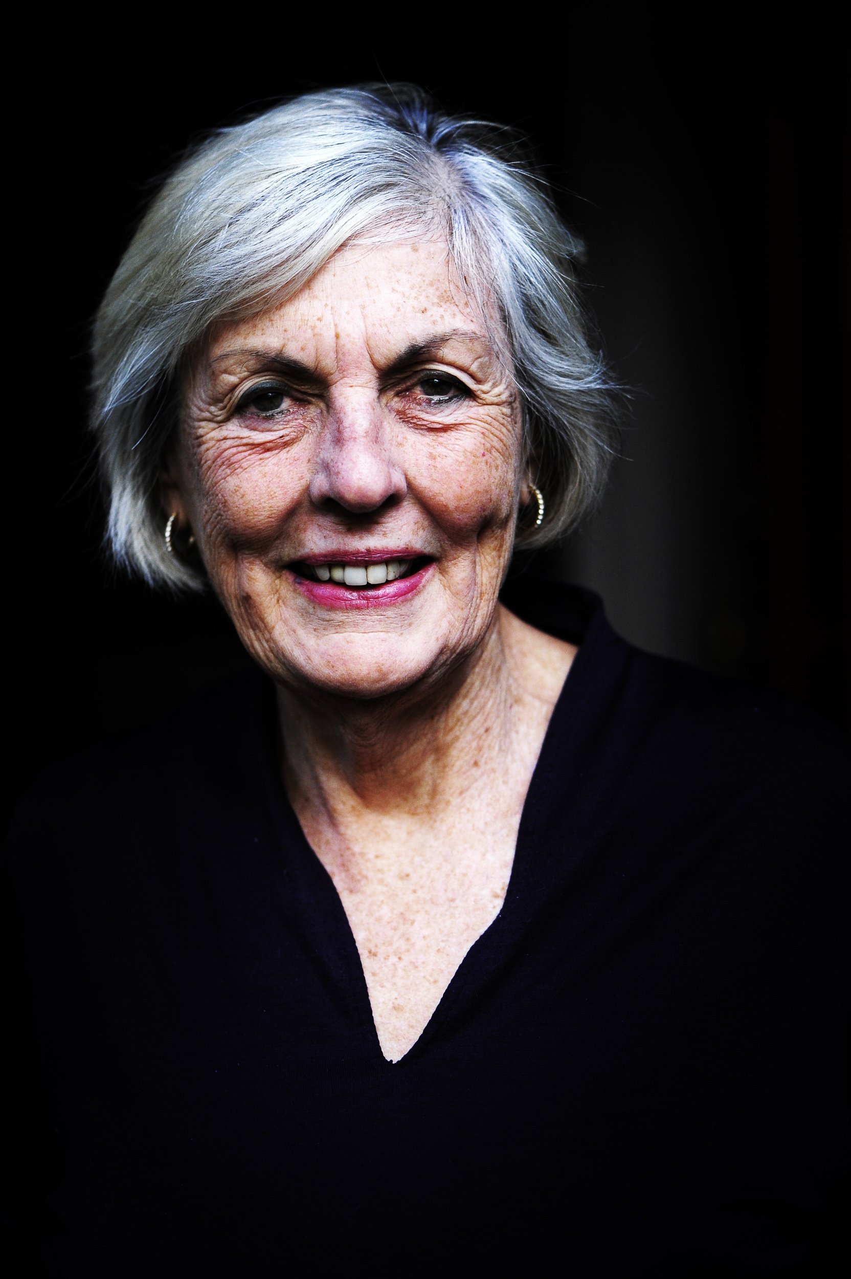An elderly woman with short gray hair, smiling, wearing a black top, with earrings, against a dark background.
