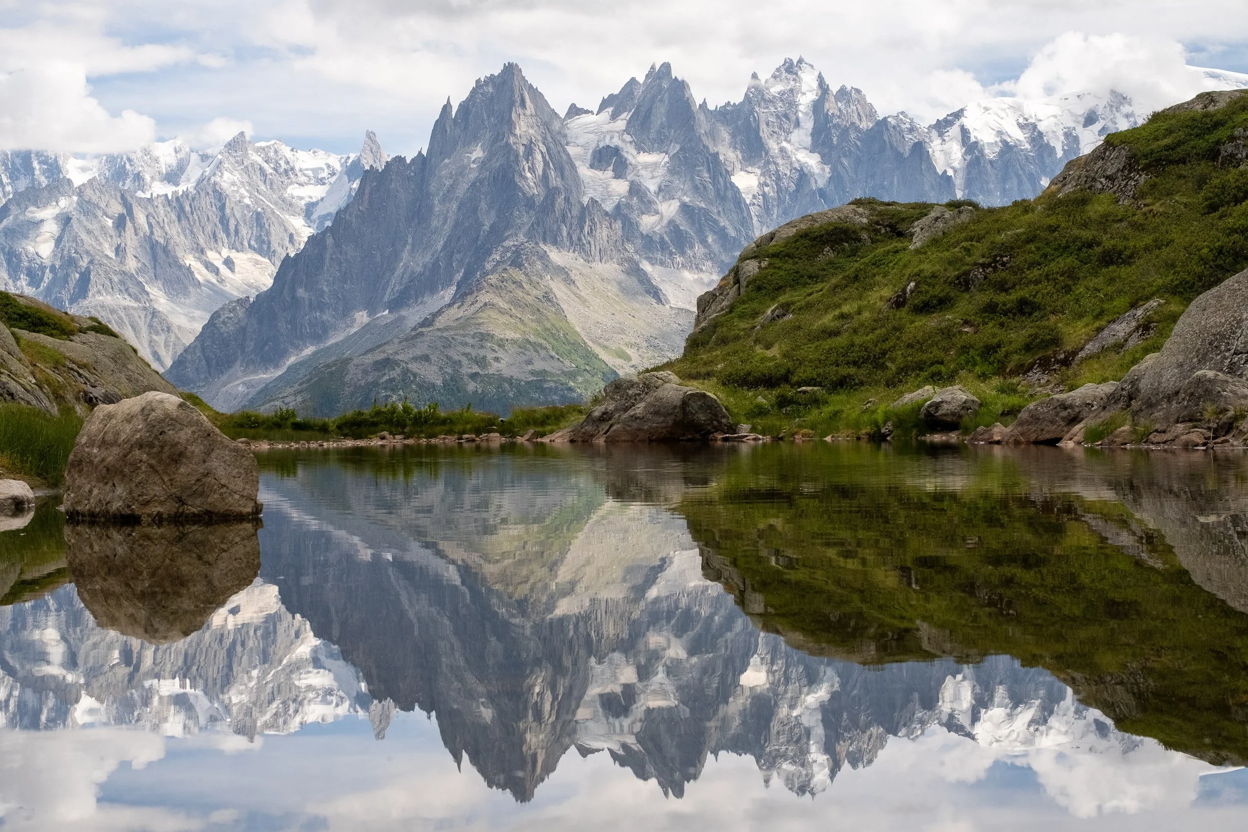 Snow-capped mountains reflecting in a peaceful lake with green hills and rocks in the foreground.