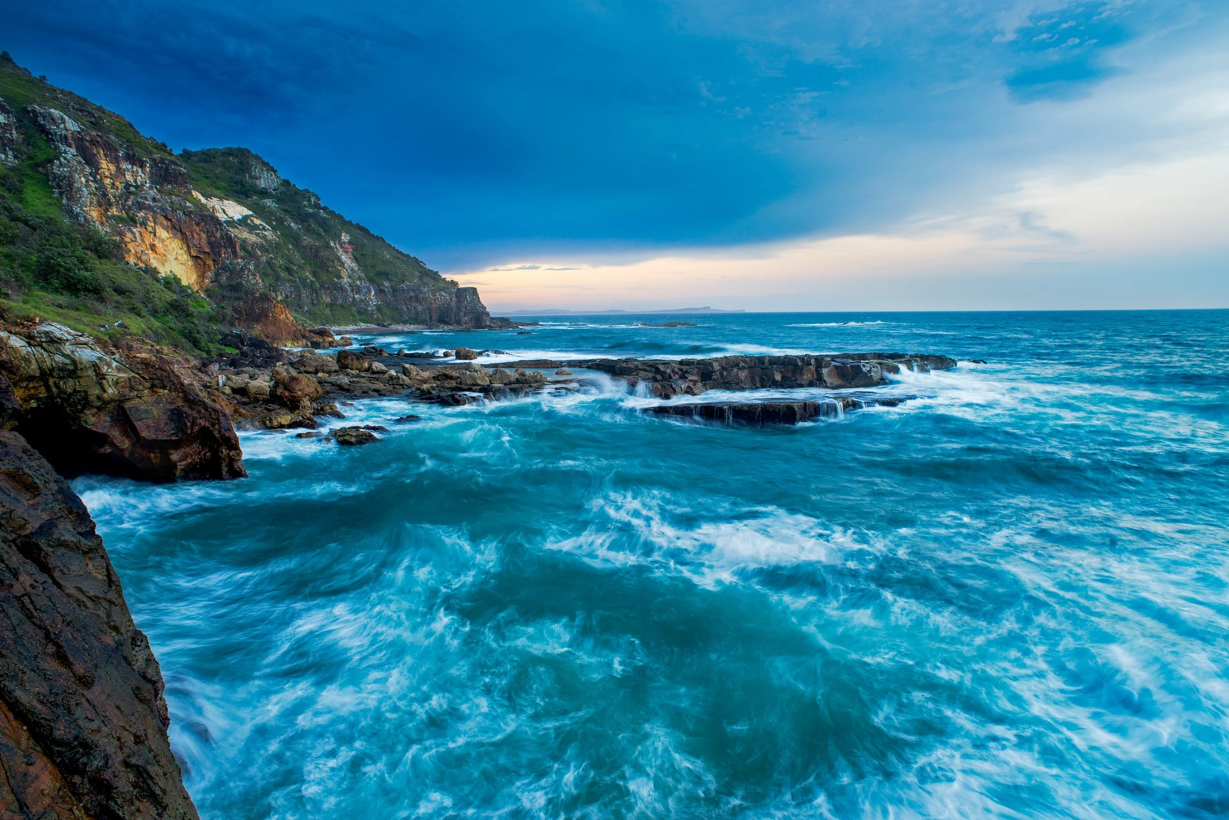 Coastline with rocky cliffs, ocean waves crashing on rocks, and a cloudy sky at sunset