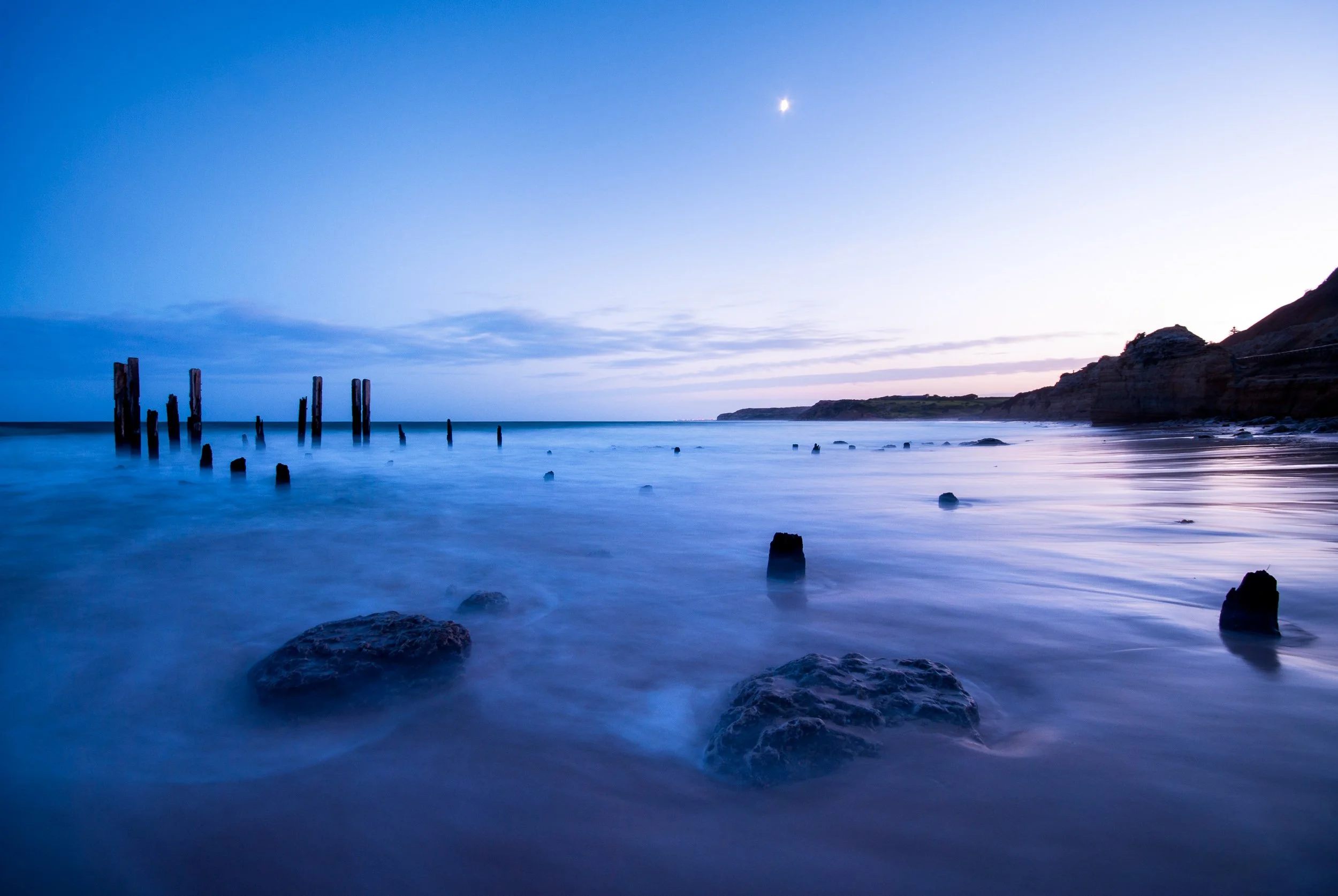 Seascape at dusk with calm waters, old wooden pilings, rocks in the foreground, and a distant cliff.