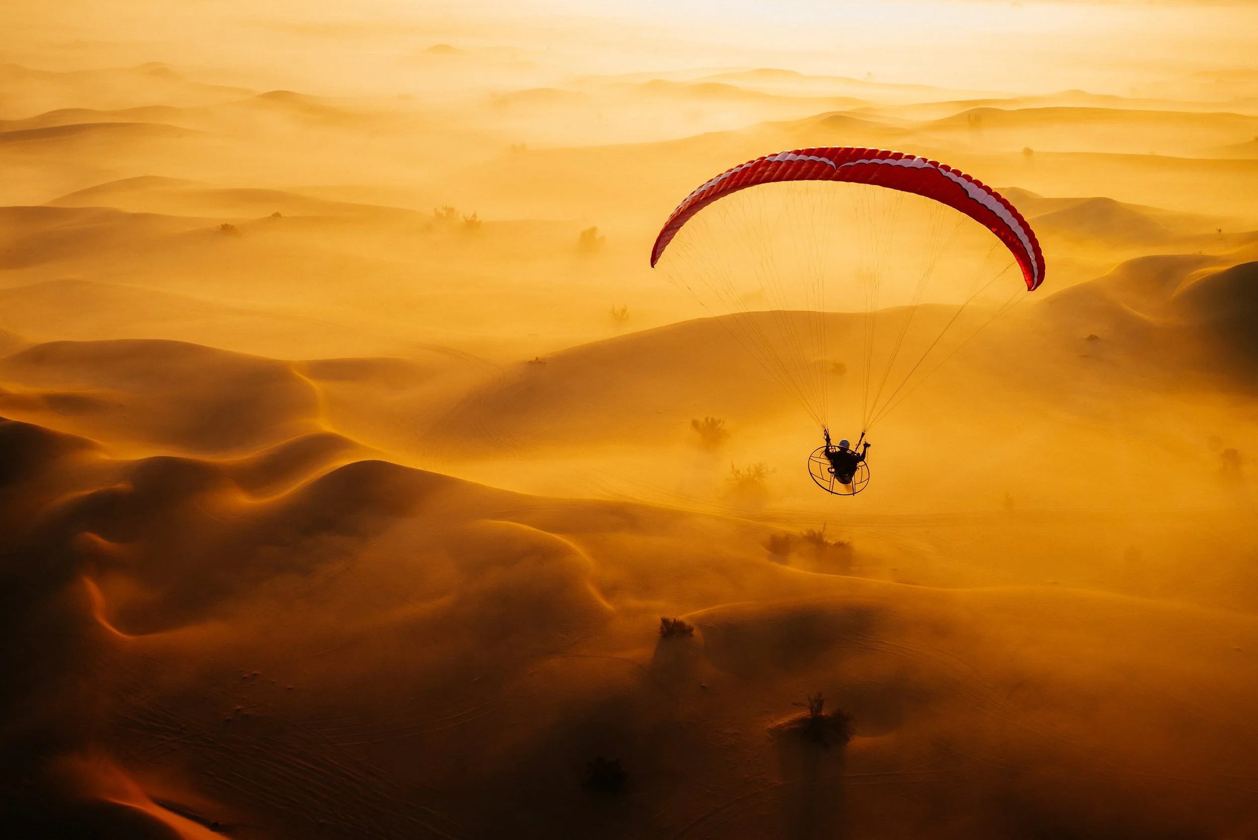 A person flying a powered paraglider over a desert landscape with sand dunes at sunset.