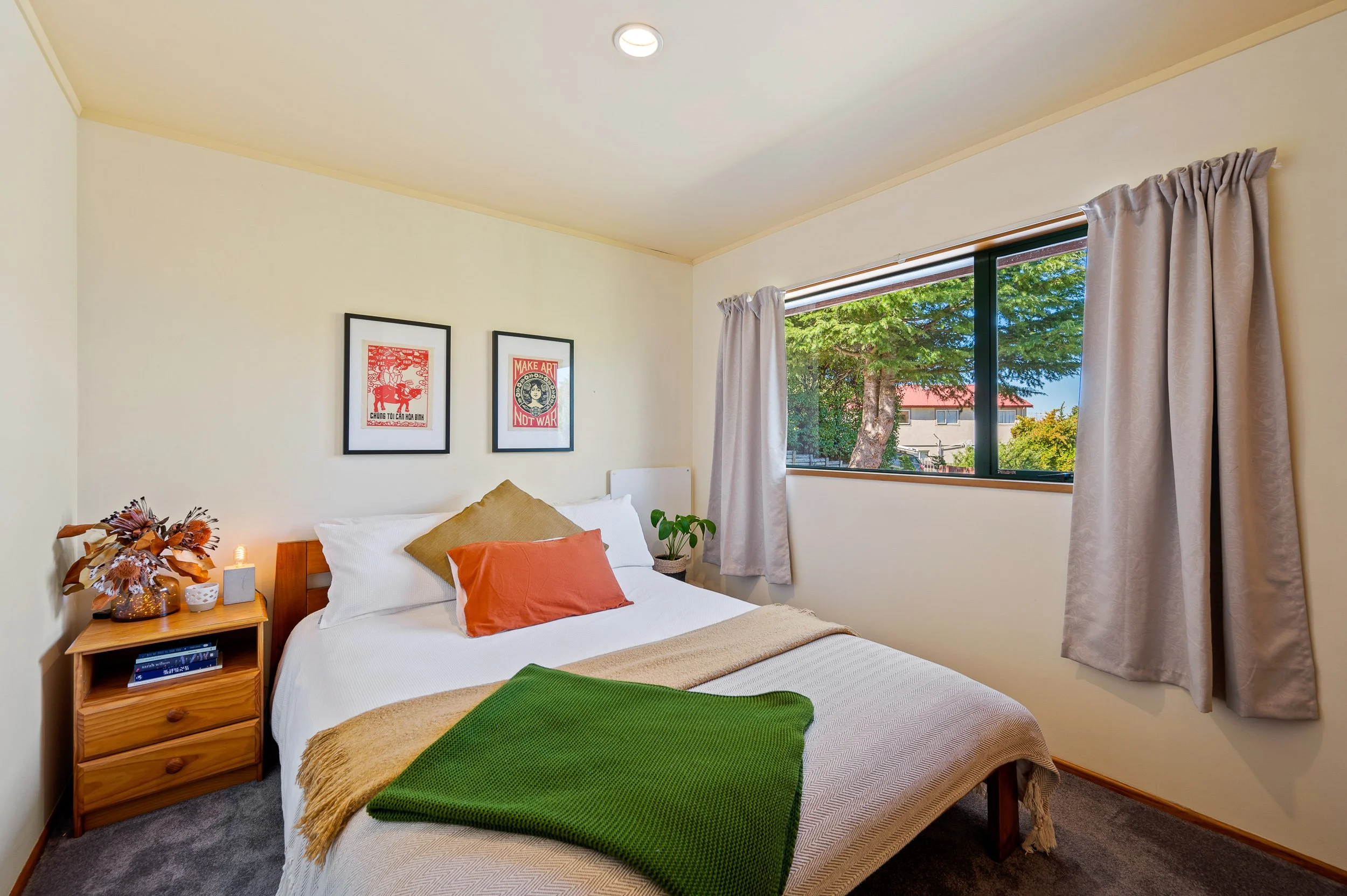 A cozy bedroom with a bed, colorful pillows, a green blanket, and a bedside table with a vase, flowers, and a lamp. There are two framed posters on the wall and a large window showing trees and neighboring buildings.