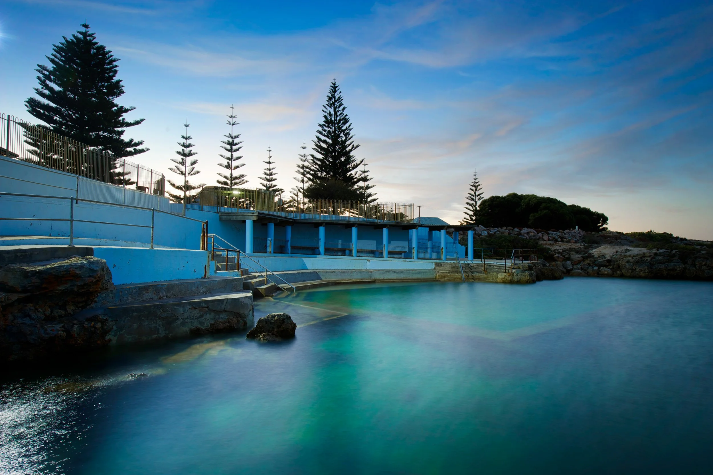 A coastal hot spring bath with a blue-painted edge, set against a rocky shoreline and several tall pine trees, during sunset or dawn with a partly cloudy sky.