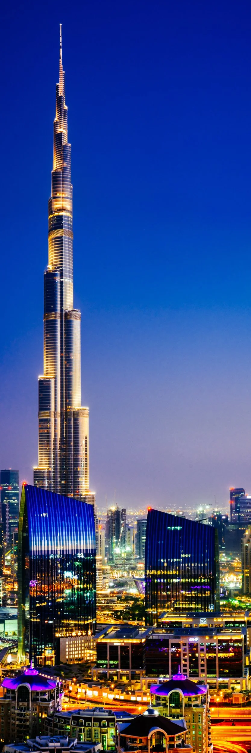 Nighttime cityscape featuring the illuminated Burj Khalifa skyscraper in Dubai, with surrounding modern buildings and bright city lights.