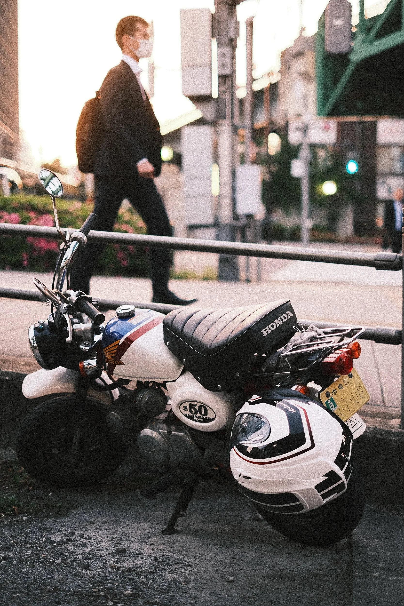 A small white Honda motorcycle with a black seat and a helmet hanging from the side, parked on a city street with a person in business attire and a face mask walking in the background.