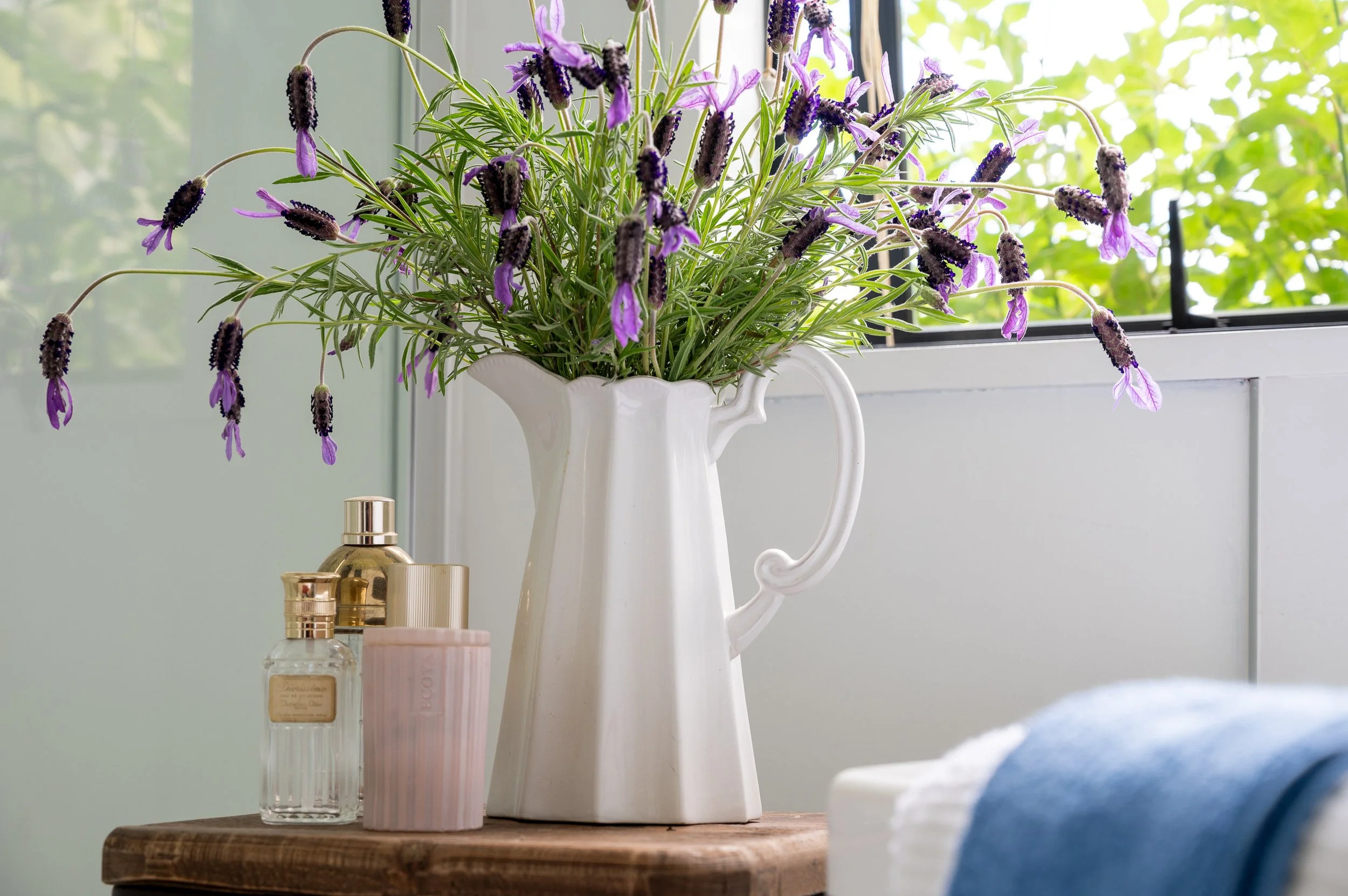 A white ceramic pitcher filled with purple lavender flowers is placed on a wooden surface near a window, with mistreated bottles and perfume containers nearby. The window shows green foliage outside.