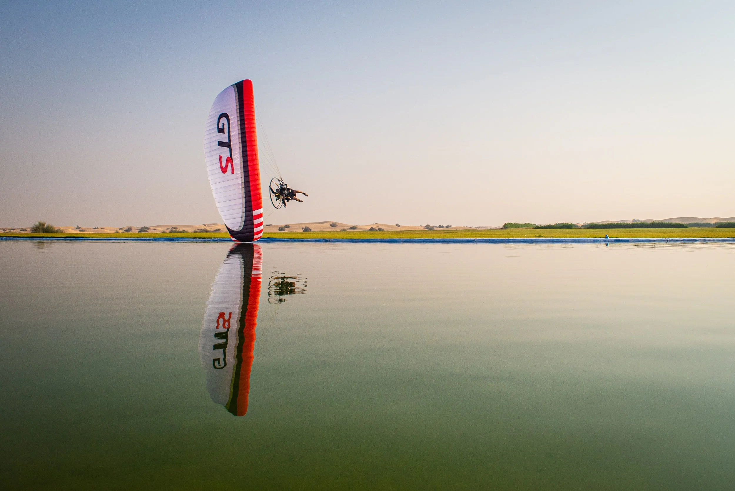 A person tandem paragliding over a body of water at sunset, with a clear sky and distant desert landscape.