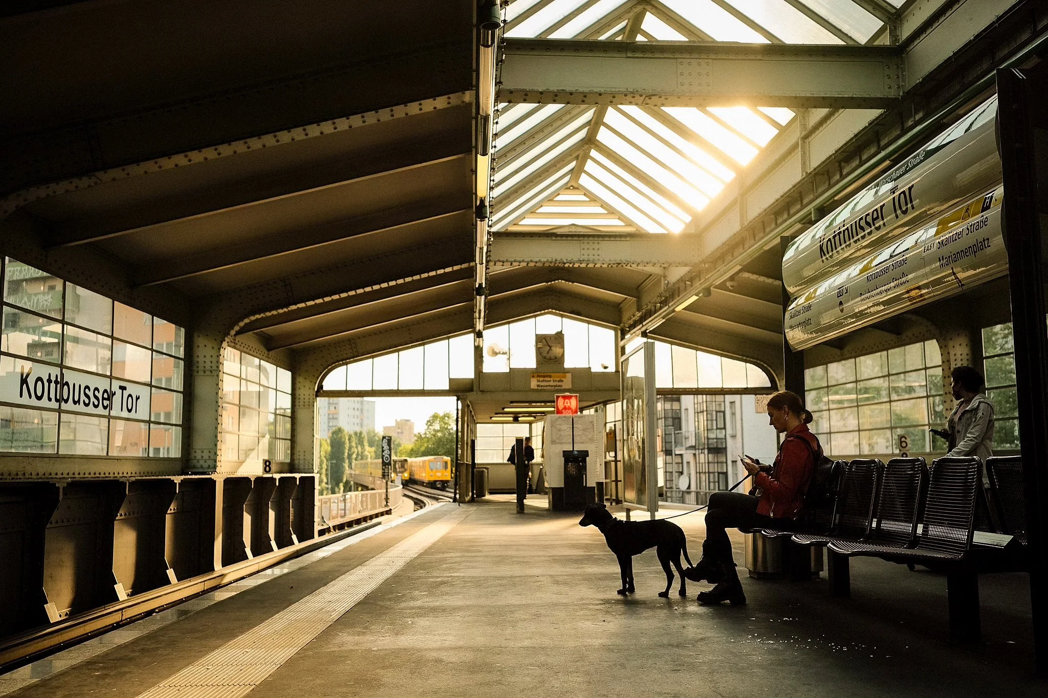 A train station platform with a woman sitting on a bench looking at her phone, a person standing near the ticket machine, and a person with a dog sitting on the bench. The platform is labeled 'Kottbusser Tor' and there is a train approaching in the b
