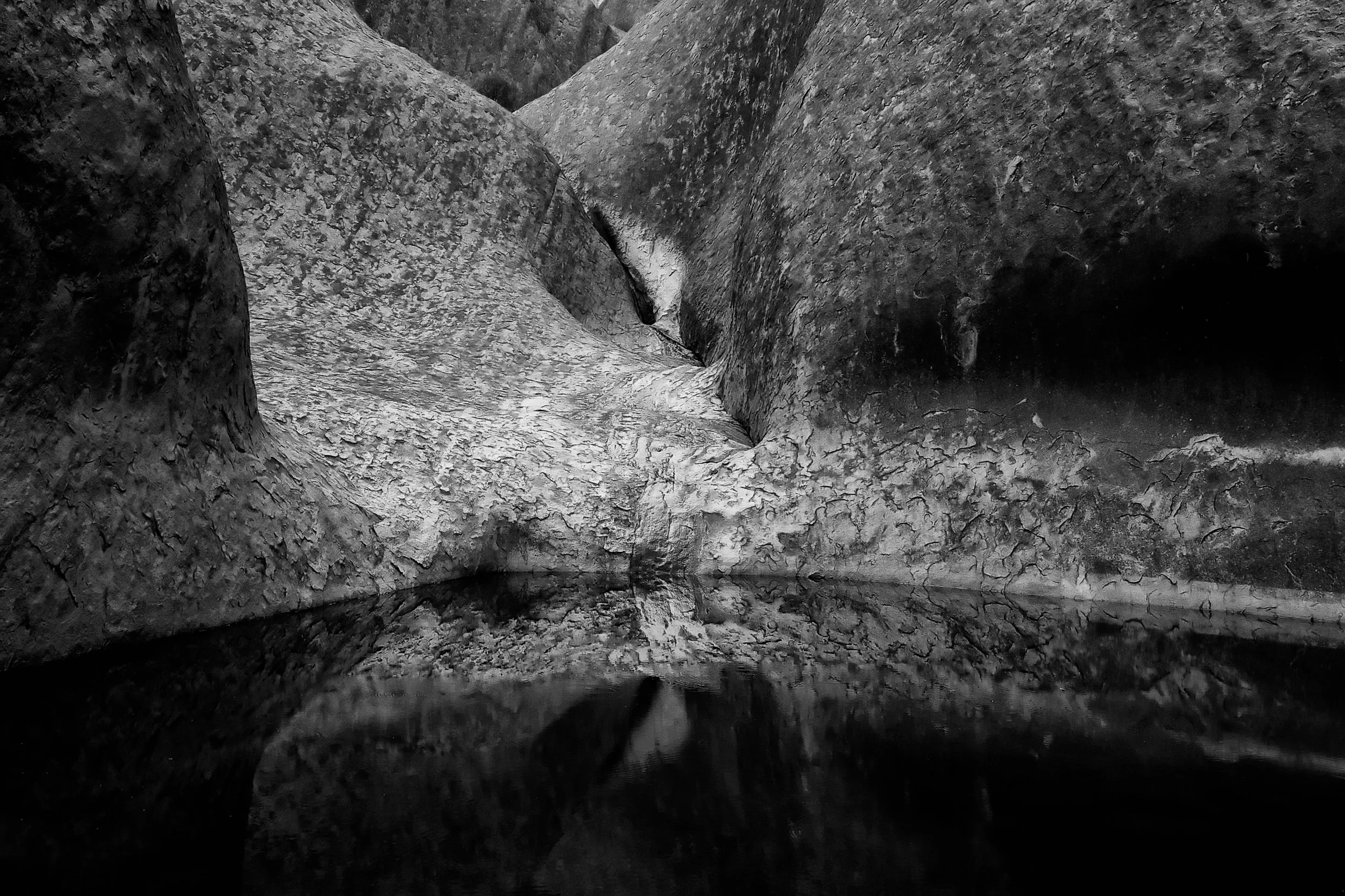Black and white photograph of a narrow canyon with steep, textured rock walls and water at the bottom reflecting the rocks.