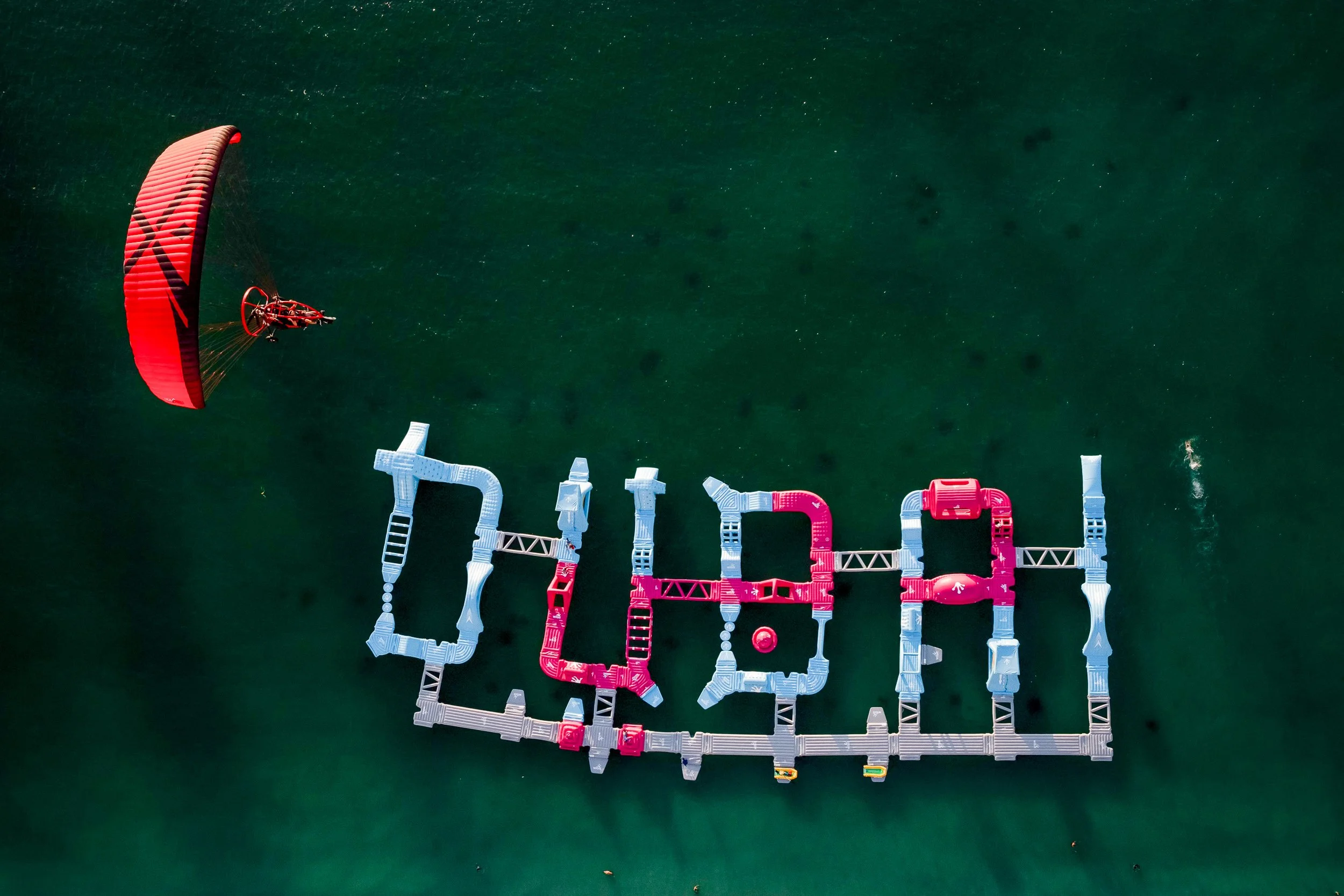 A floating dock in the water spells out the word "DREAM" with colorful modular sections, and a paramotor with a red wing is flying nearby.