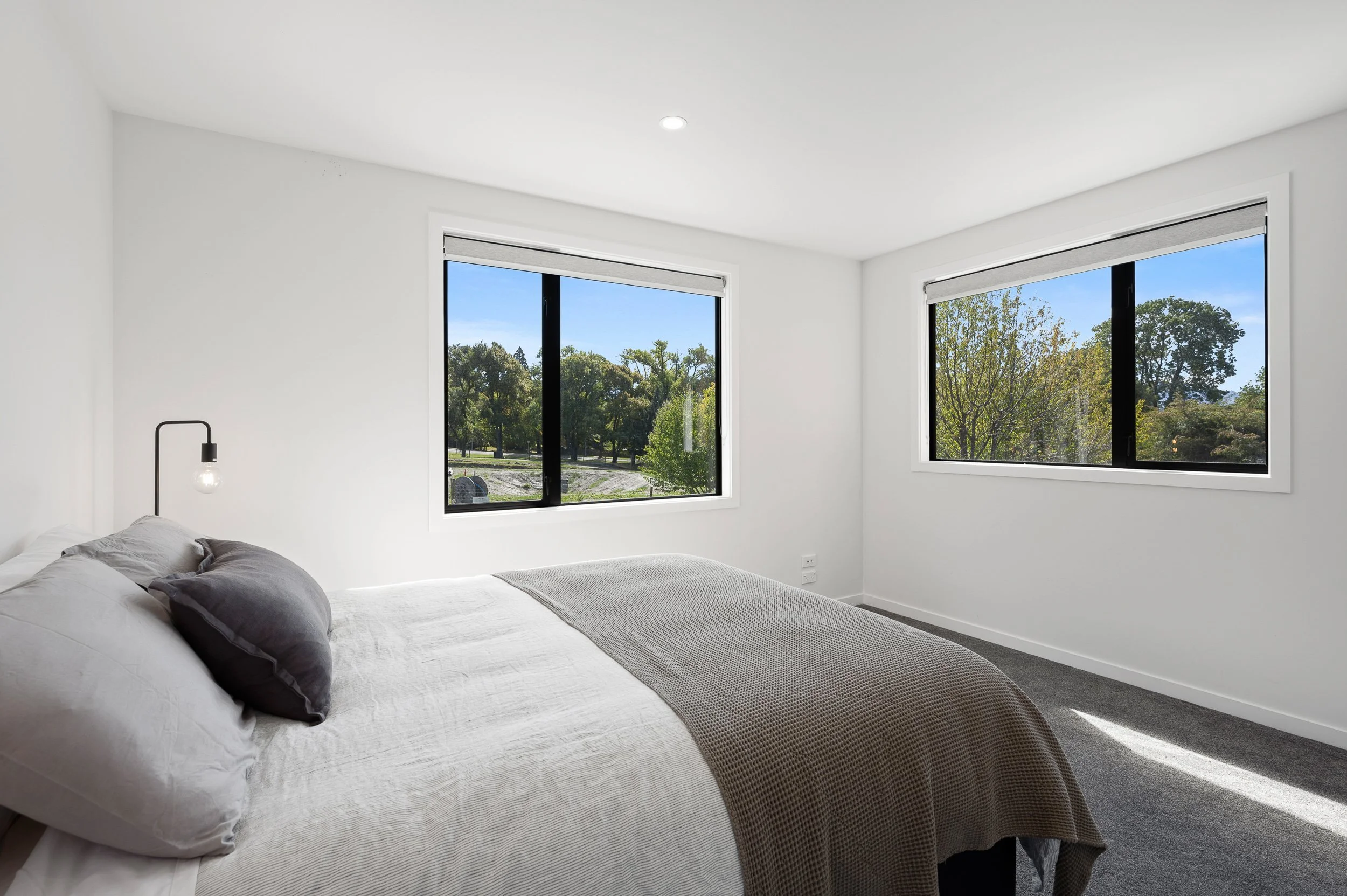 A minimalist bedroom with white walls, a bed with white and gray bedding, and two large windows showing a view of trees and blue sky.