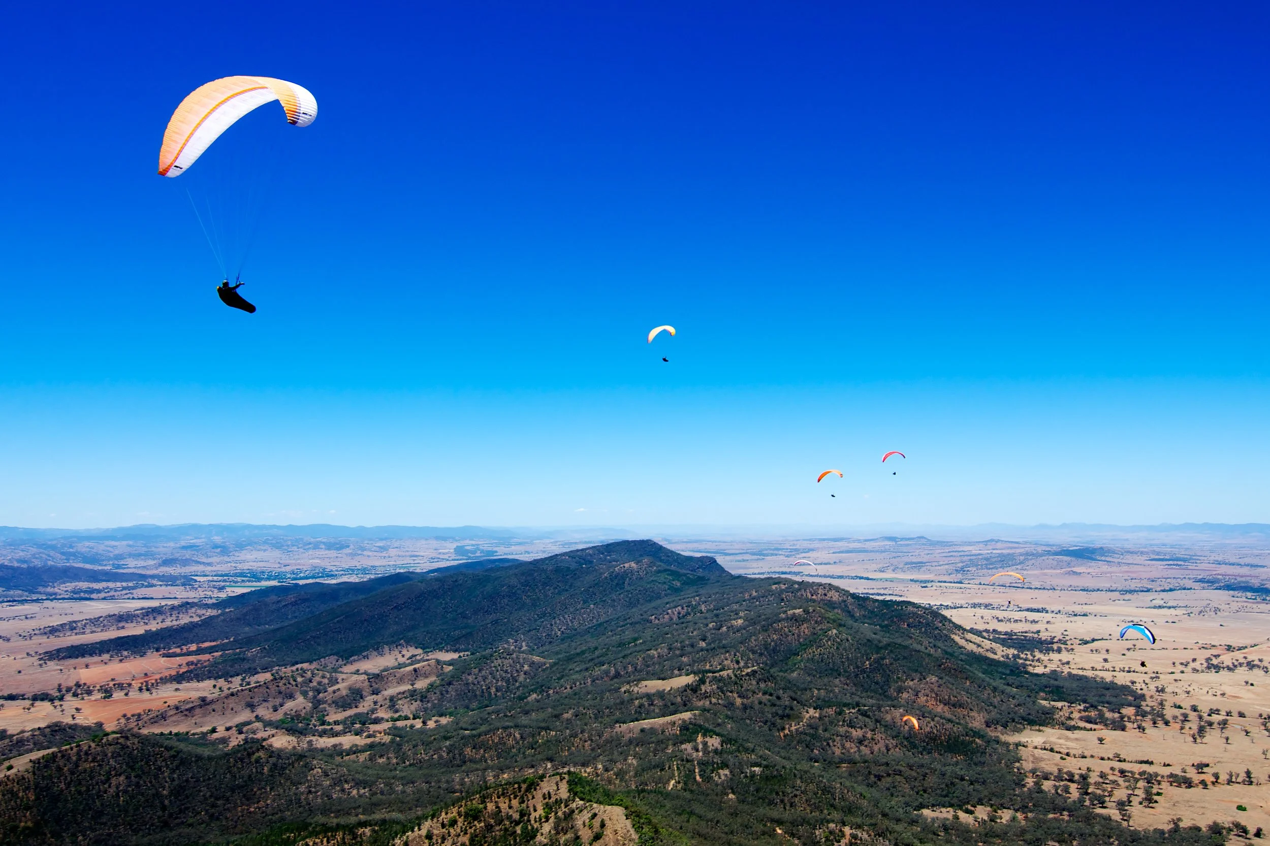 Multiple paragliders flying over a landscape of hills and plains under a clear blue sky.