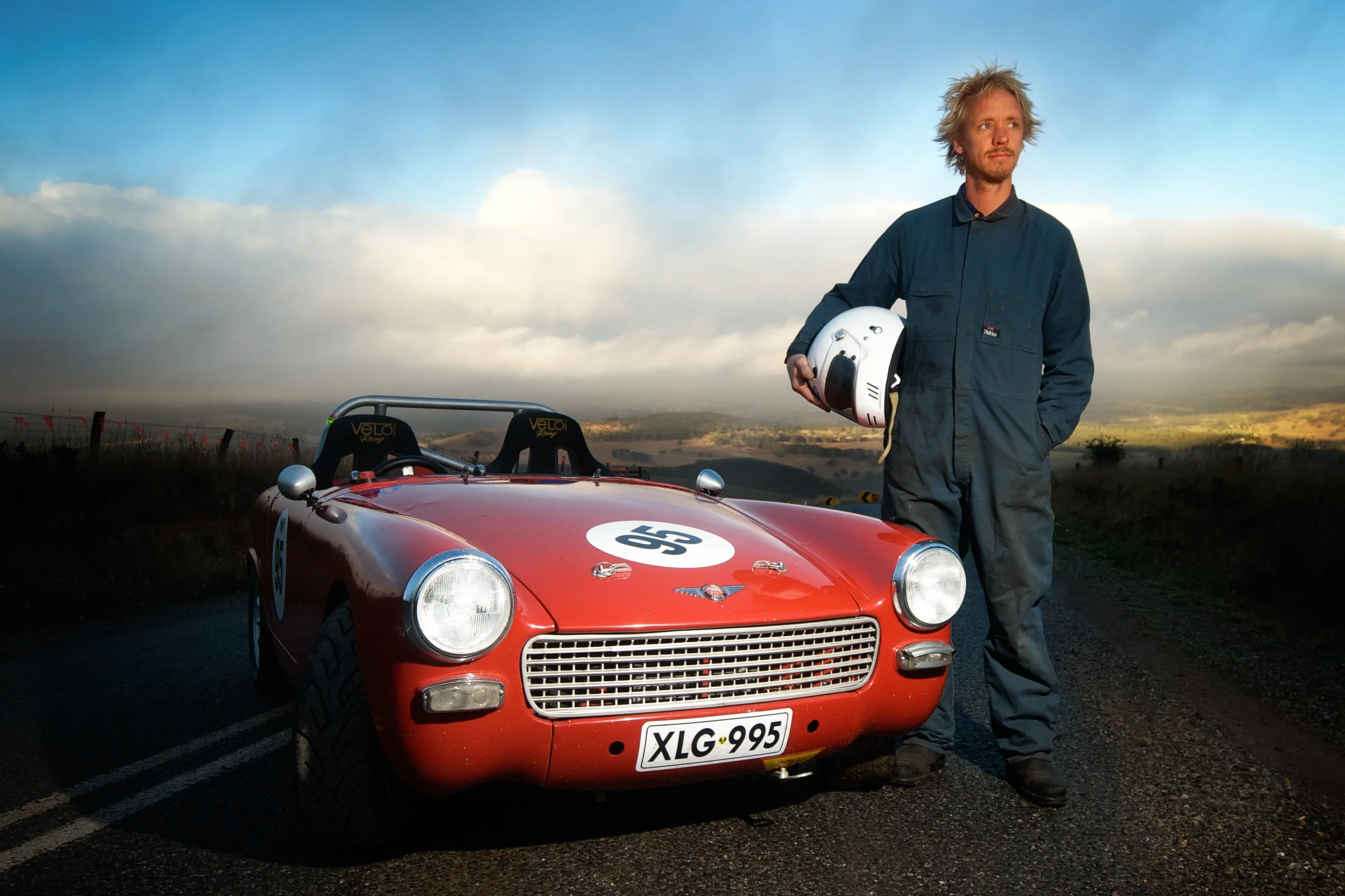 A man standing next to a vintage red race car holding a white helmet, on a rural road with an open landscape and cloudy sky in the background.