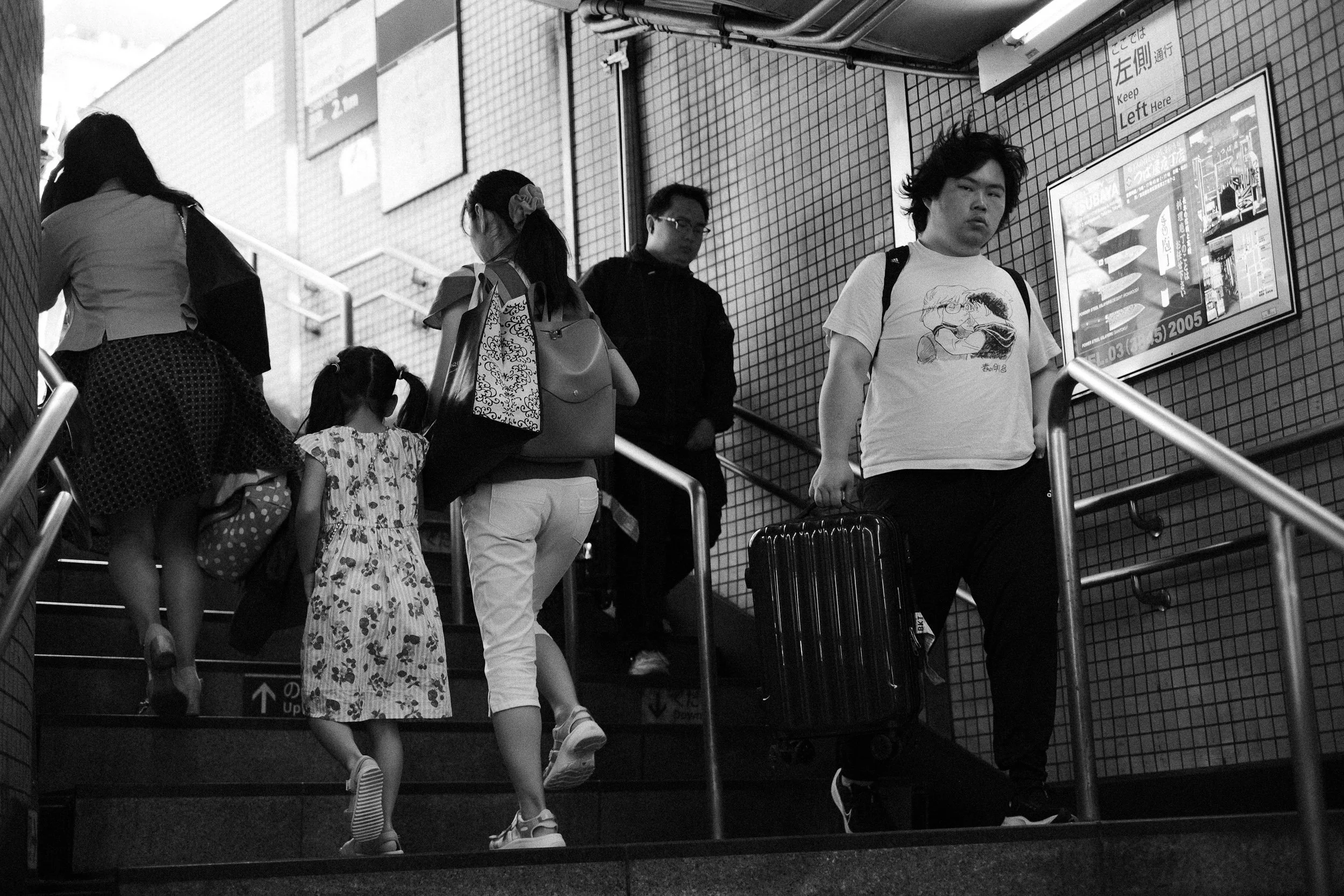 People ascending a stairway in a public area, with some carrying luggage and bags, and a woman with a young girl holding hands. The setting appears to be indoors, possibly a transit station or shopping mall.