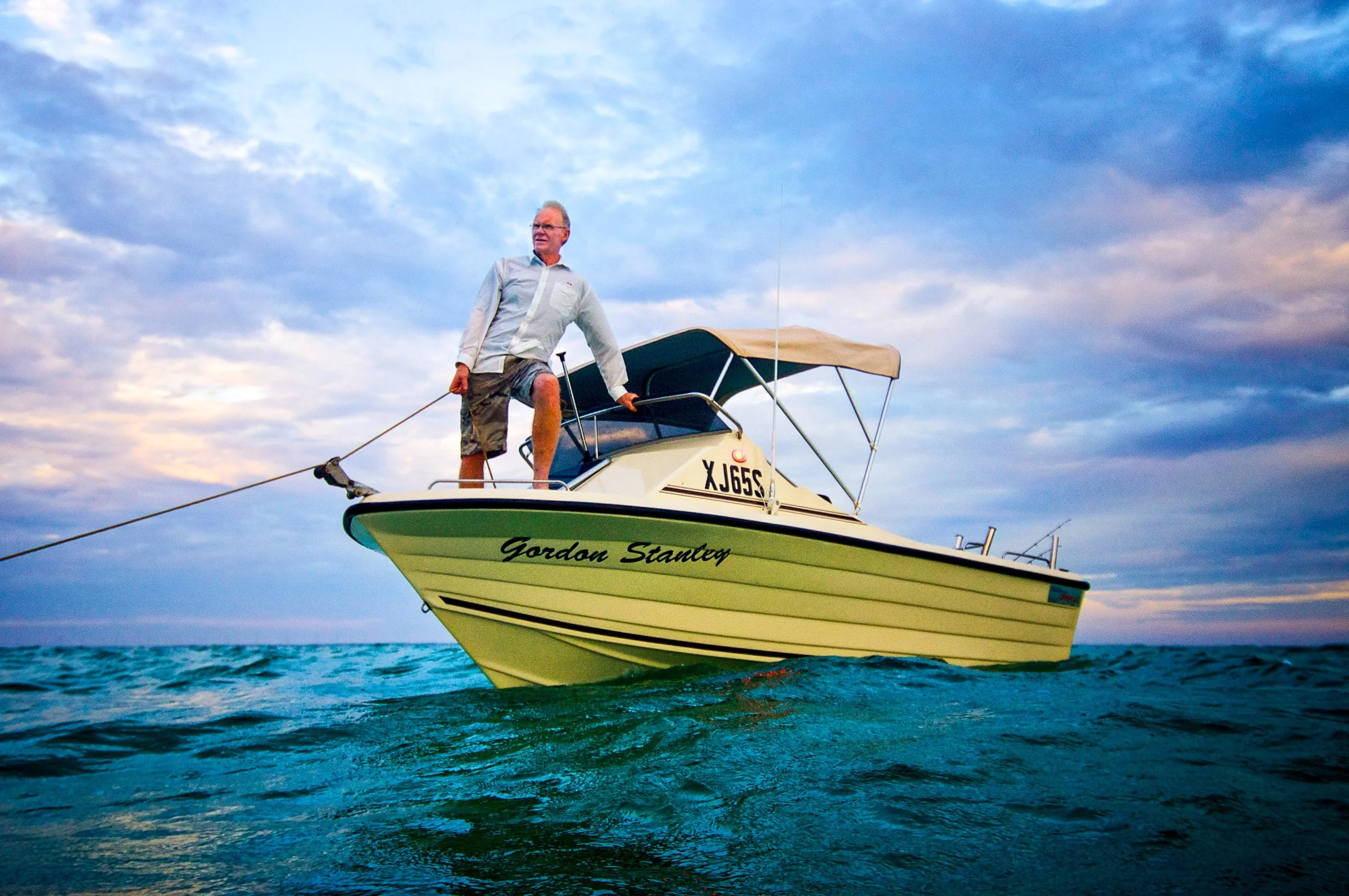 A man standing on a boat named 'Gordon Stanteg' on the water, holding a rope, with a cloudy sky in the background.