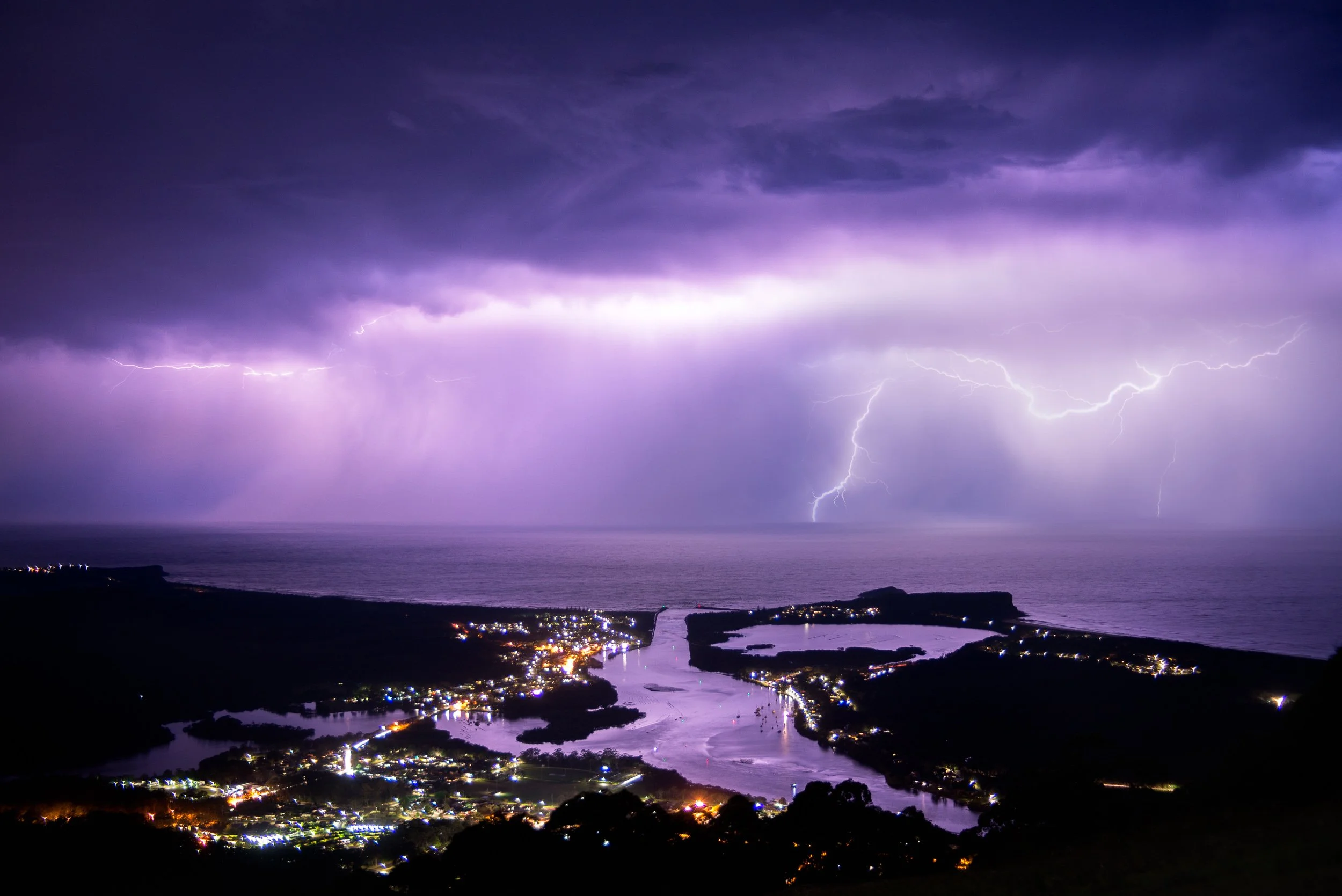 Lightning storm over a coastal town at night, with dark clouds, illuminated by city lights and lightning strikes across the sky.