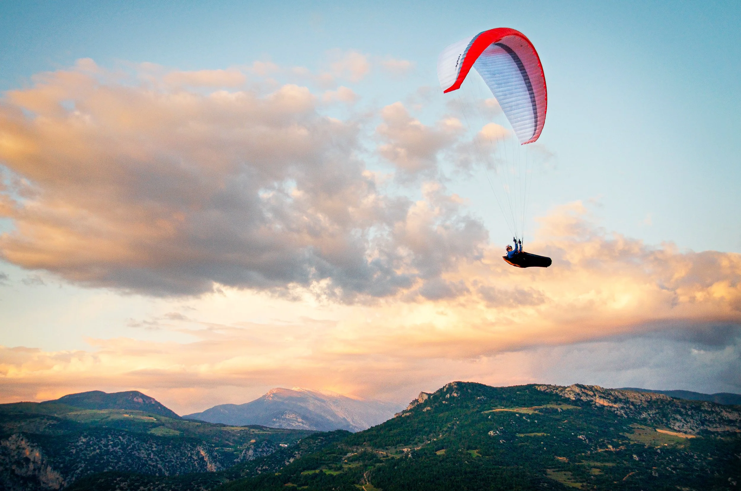 A person paragliding over a mountainous landscape during sunset with a red and white parachute.