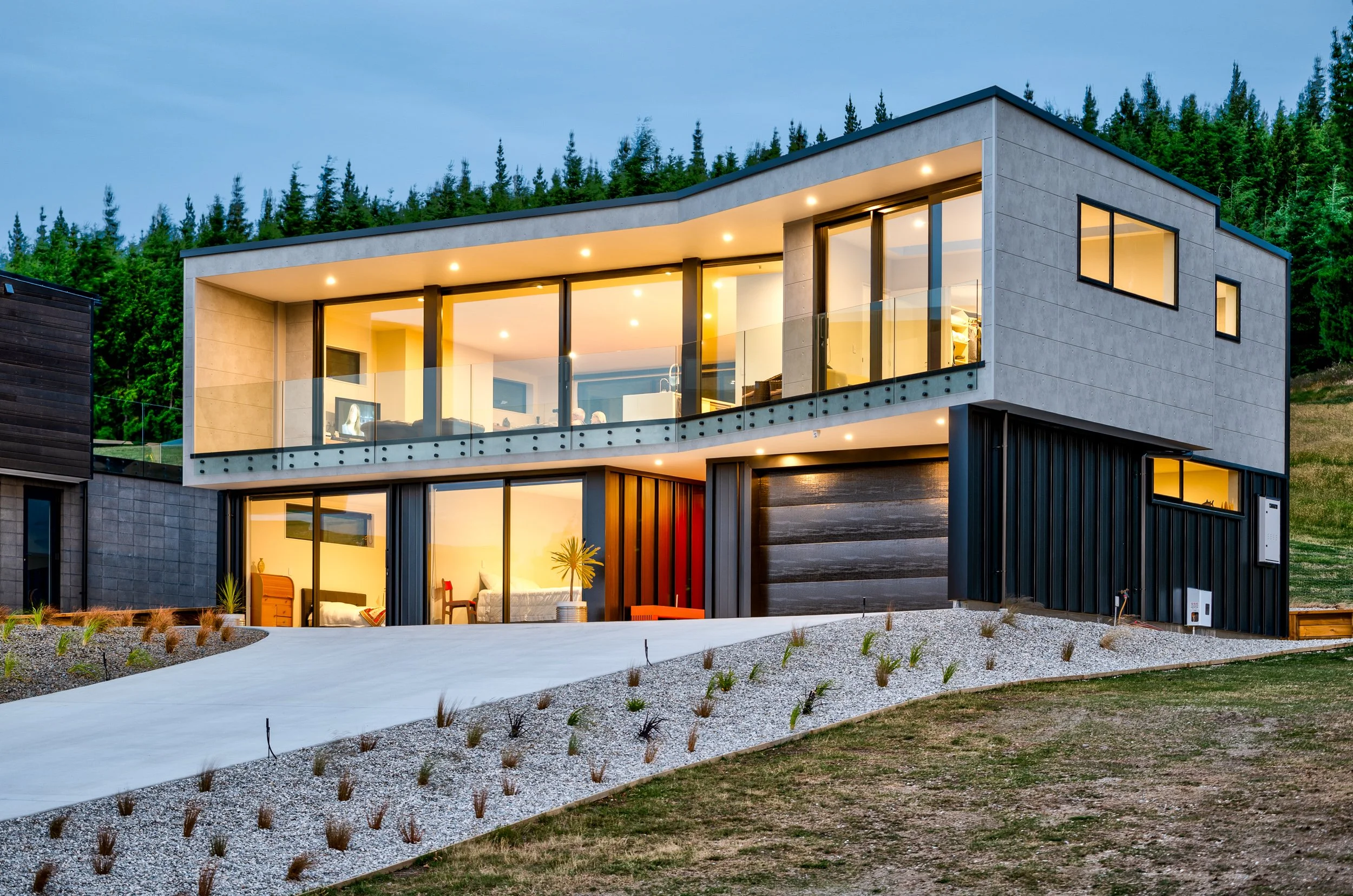 Modern two-story house with large glass windows and a balcony, surrounded by outdoor landscaping and forested hills in the background, illuminated at dusk.