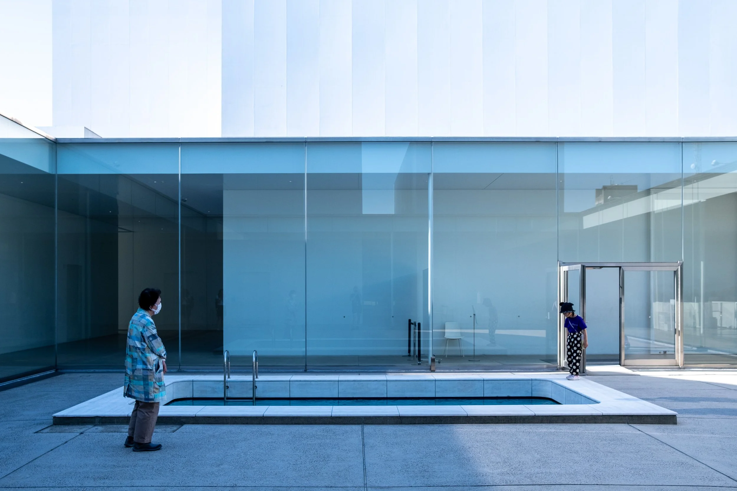 A modern building with large glass windows. Two people wearing masks, one inside near the entrance and the other standing outside.