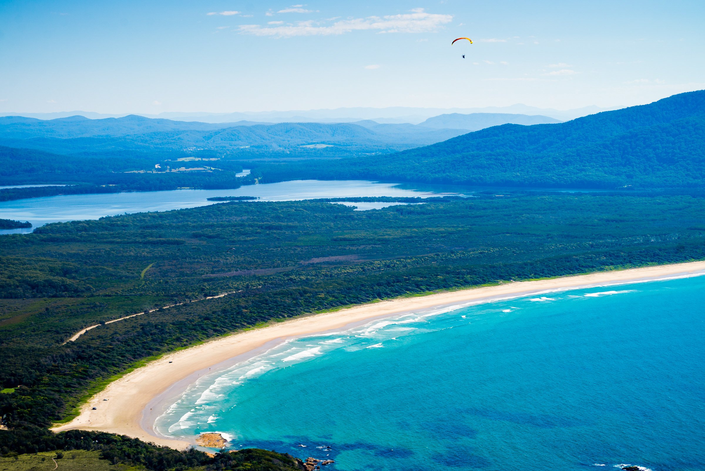 Aerial view of a beach with white sand and turquoise water, bordered by lush green forest, with hills and mountains in the background and a paraglider in the sky.