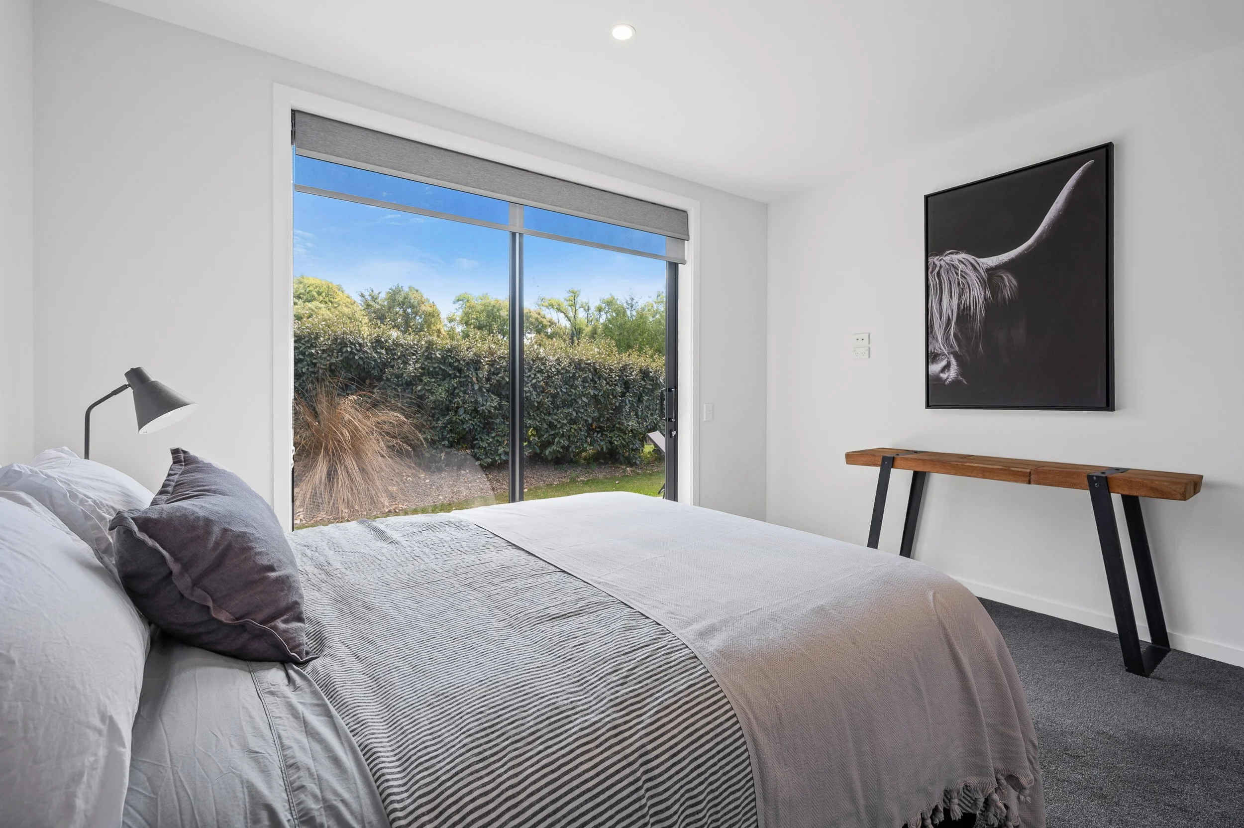 Modern bedroom with large sliding glass door facing garden, white walls, gray bedding, black and gray pillow, black wall art of a highland cow, wooden console table with black legs.