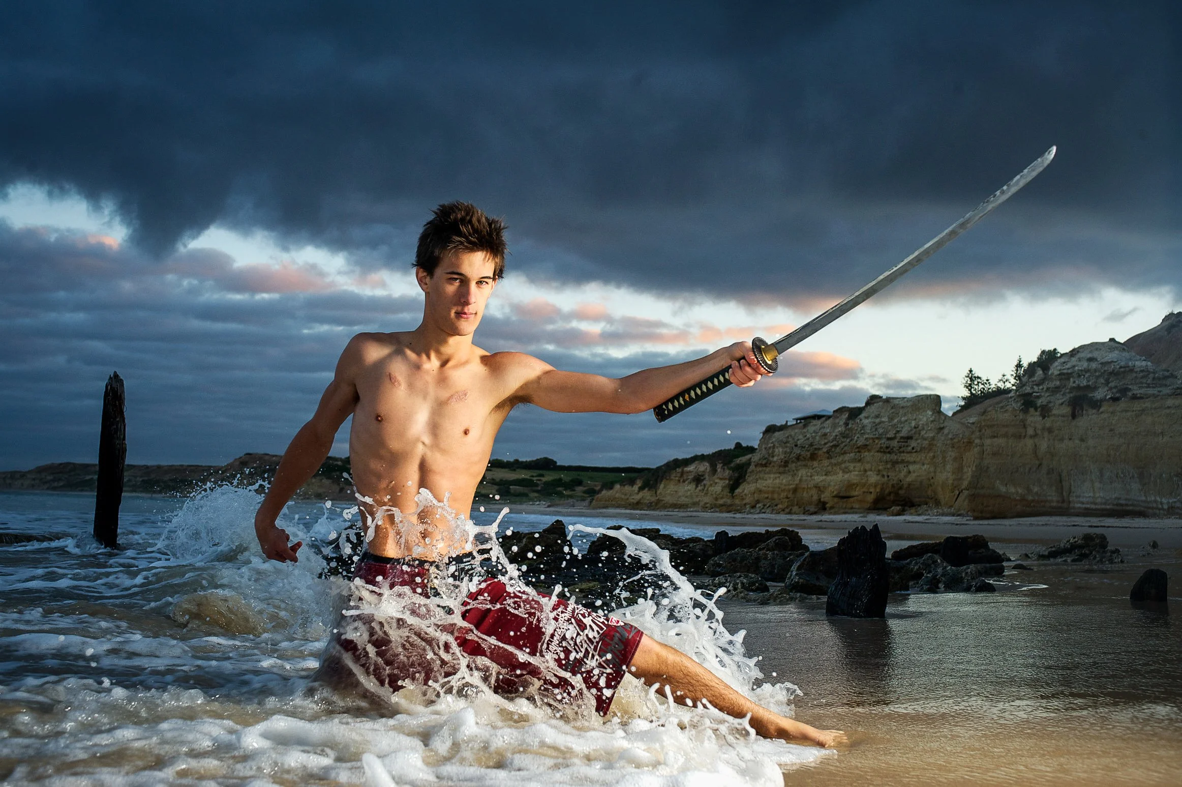 A young man with dark hair and a lean physique, shirtless and wearing red shorts, wades through the ocean waters at the beach while holding a katana sword extended outward against a backdrop of cloudy sky and rocky cliffs.