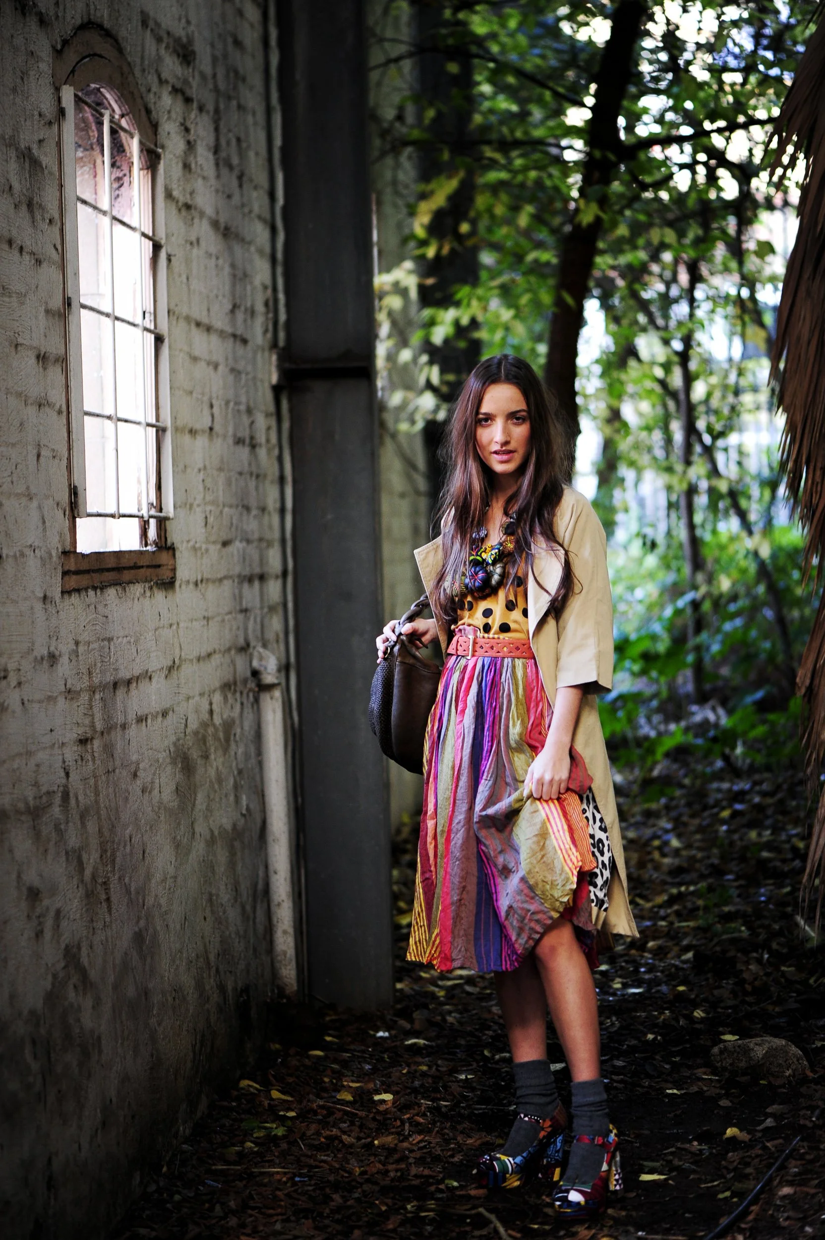 A woman with long brown hair wearing a colorful dress, beige coat, and high heels walking outdoors on a dirt path with greenery and trees in the background.