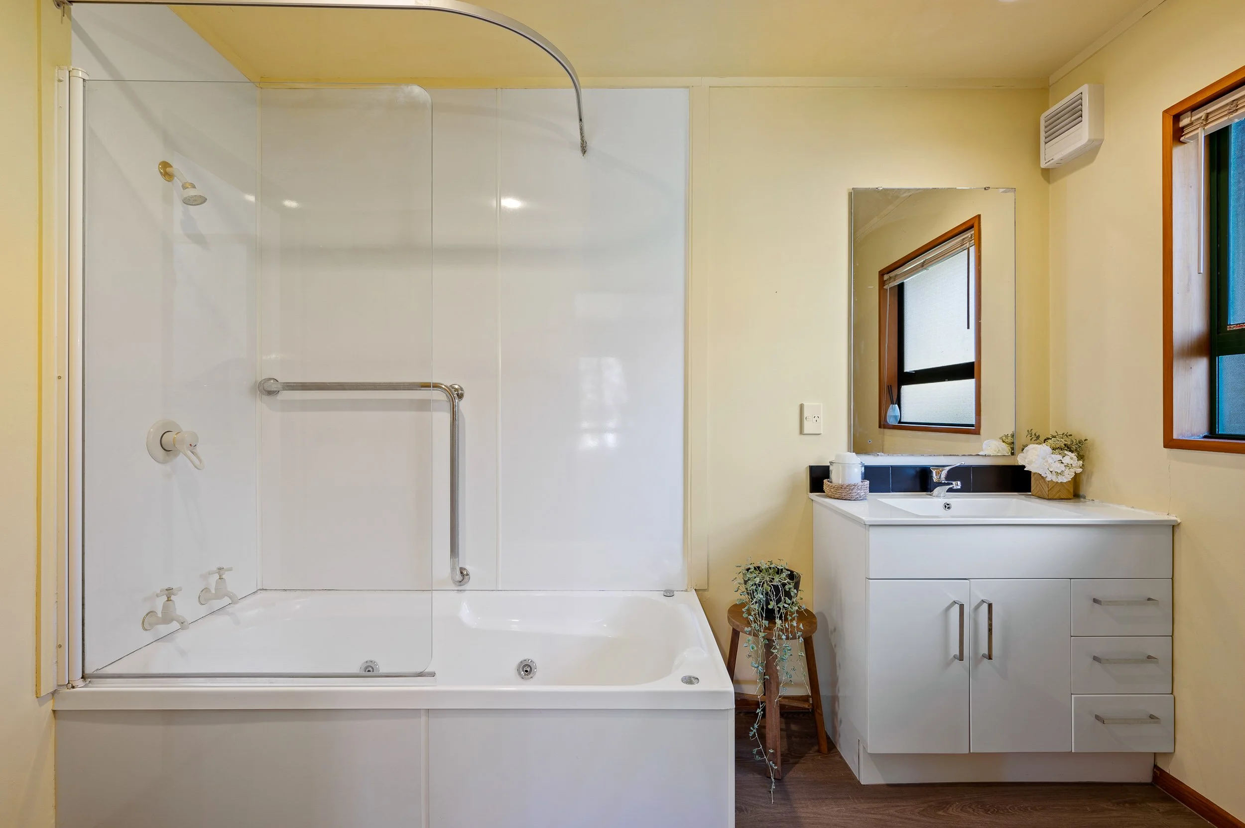 A bathroom with a white bathtub and shower on the left, and a white vanity with a mirror above and a window to the right. There is a small wooden stool with a plant on it next to the vanity, and a window with a wooden frame to the far right.