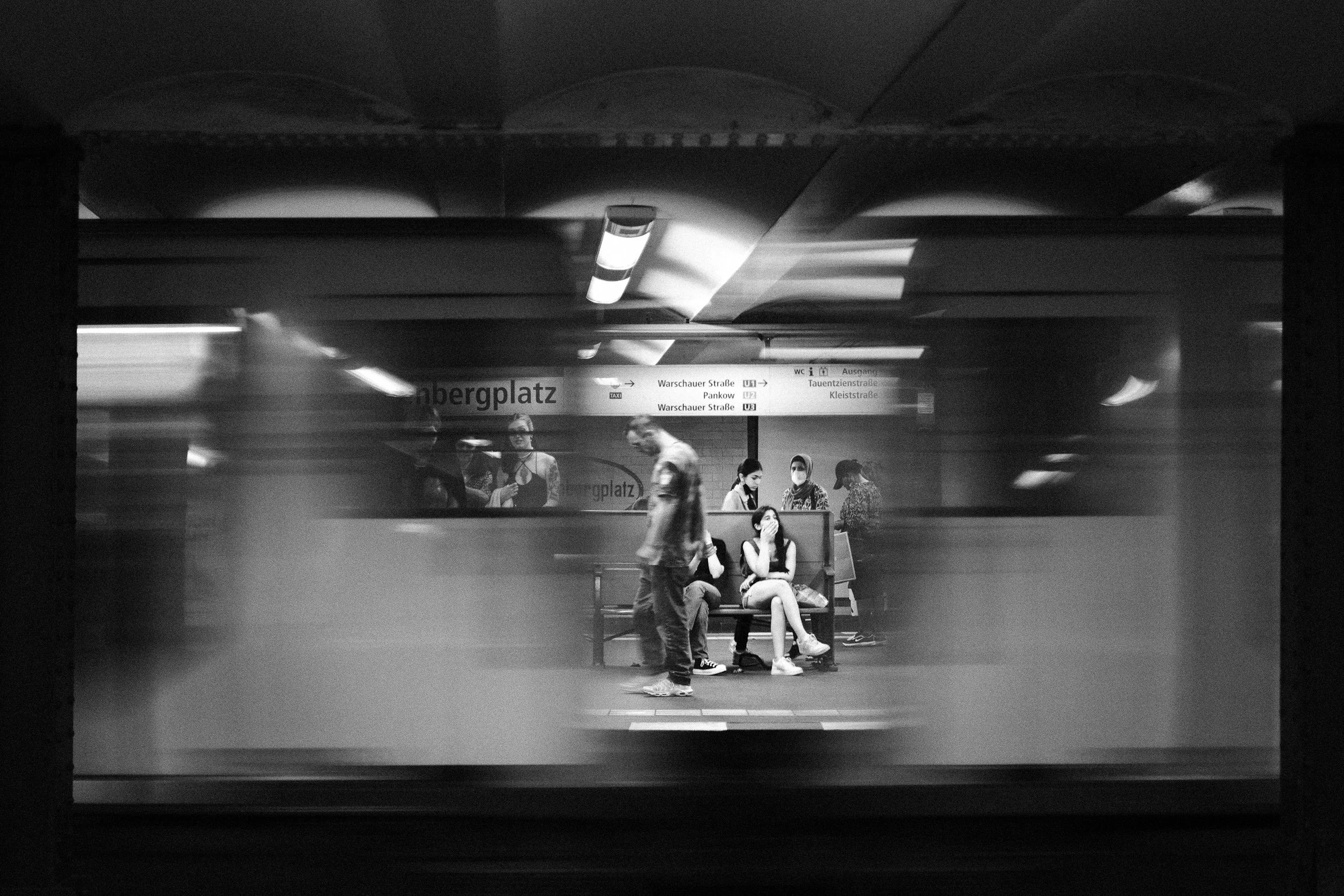 People waiting at a subway station, some sitting on a bench and others walking, with train in motion blurred behind them, in black and white.