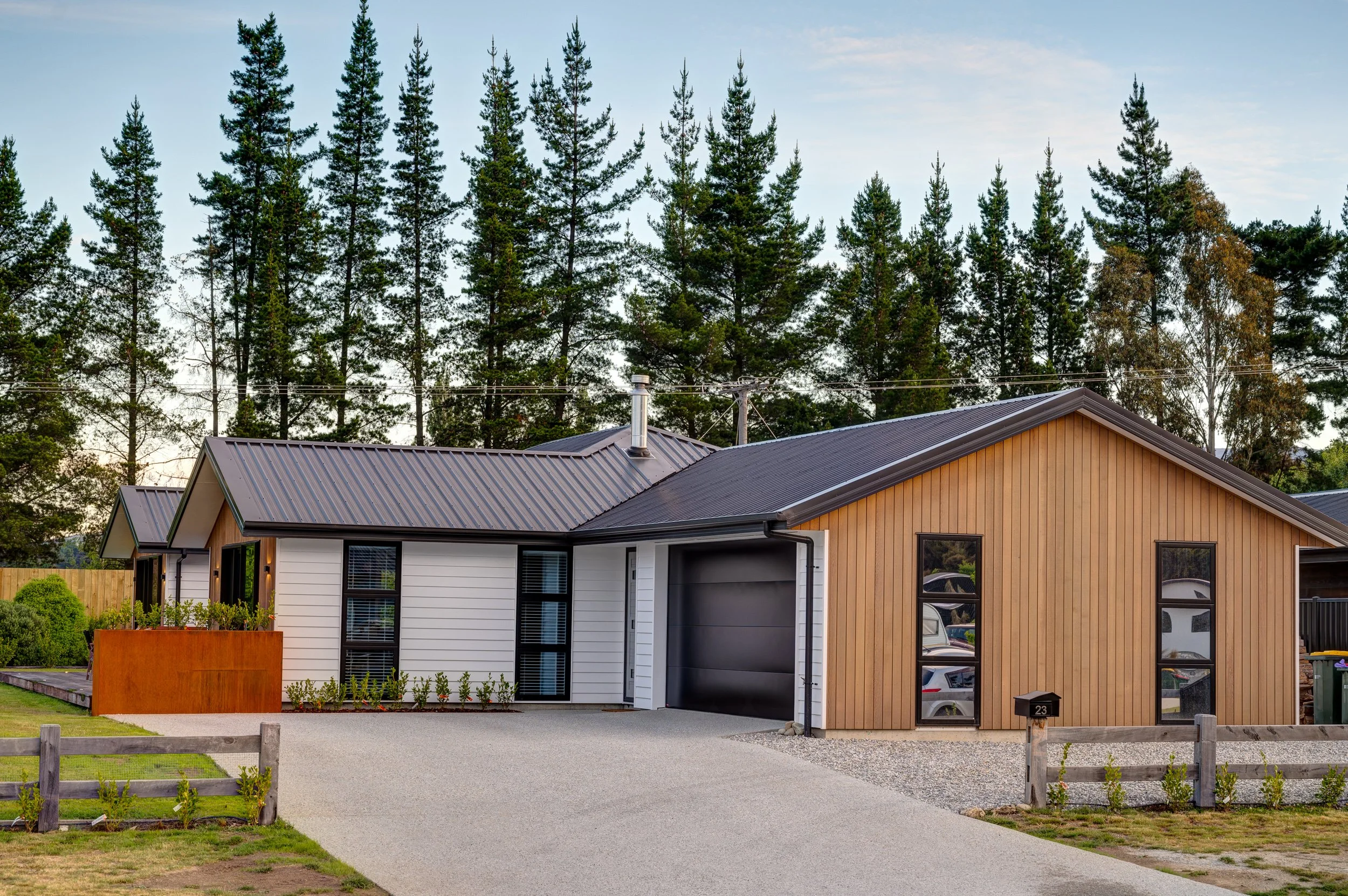 Modern house with a black garage door, wood and white siding, black-framed windows, and a driveway, set against a background of tall pine trees.