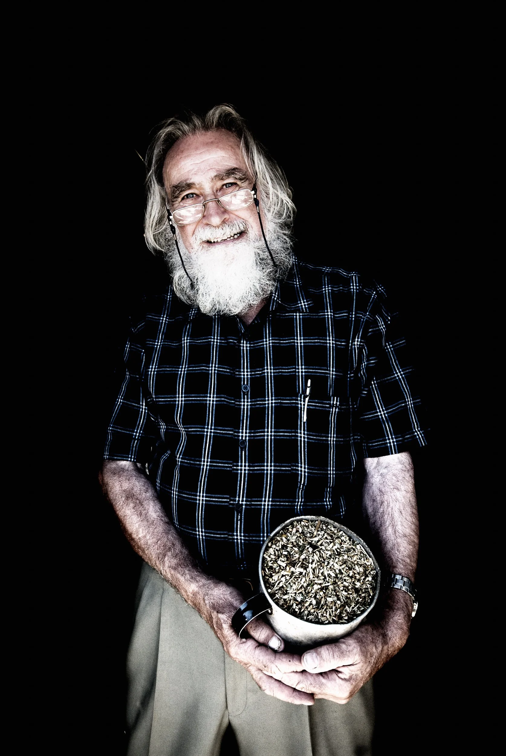 An elderly man with long white hair and a beard smiling, wearing glasses, a black and blue plaid shirt, holding a bowl filled with seeds against a black background.
