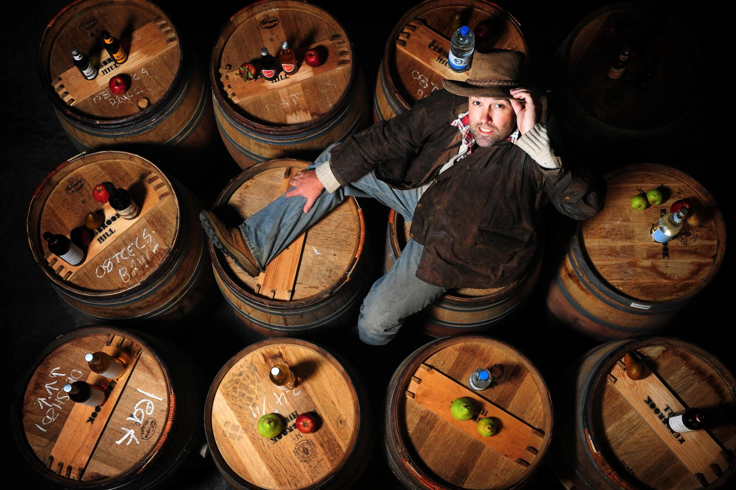 A man sitting on a barrel in a cellar surrounded by wooden barrels, some with bottles, apples, and limes on top. The man is wearing a cowboy hat, a plaid shirt, jeans, and a brown jacket.