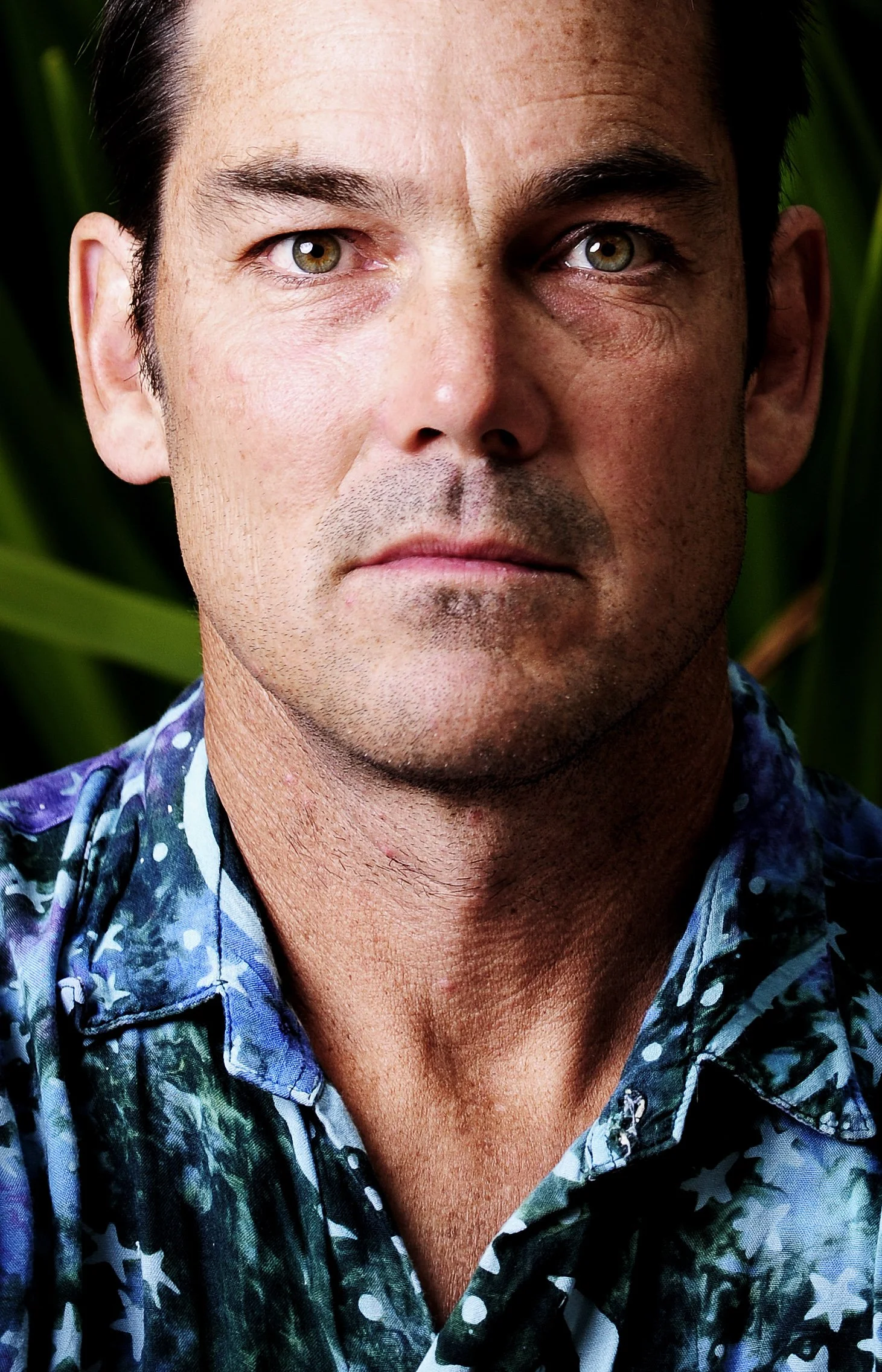 Close-up portrait of a man with short dark hair and light-colored eyes, wearing a colorful, patterned shirt. Background appears to be green foliage.