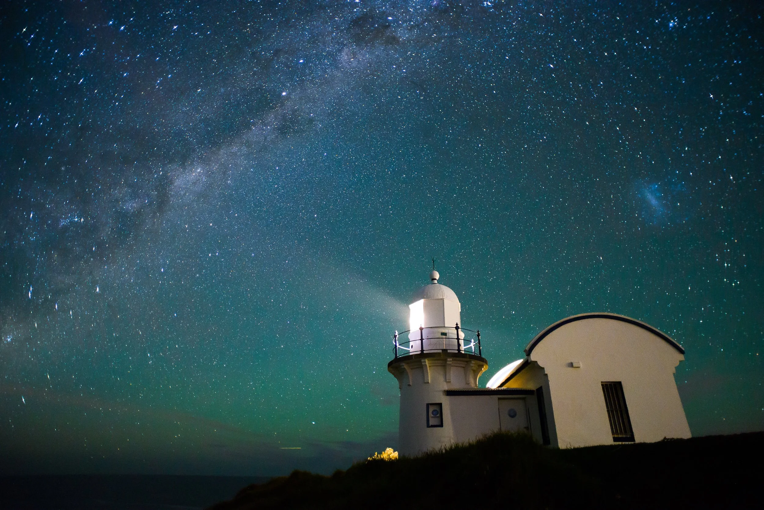 A lighthouse on a grassy hill at night with a star-filled sky and the Milky Way galaxy visible overhead.