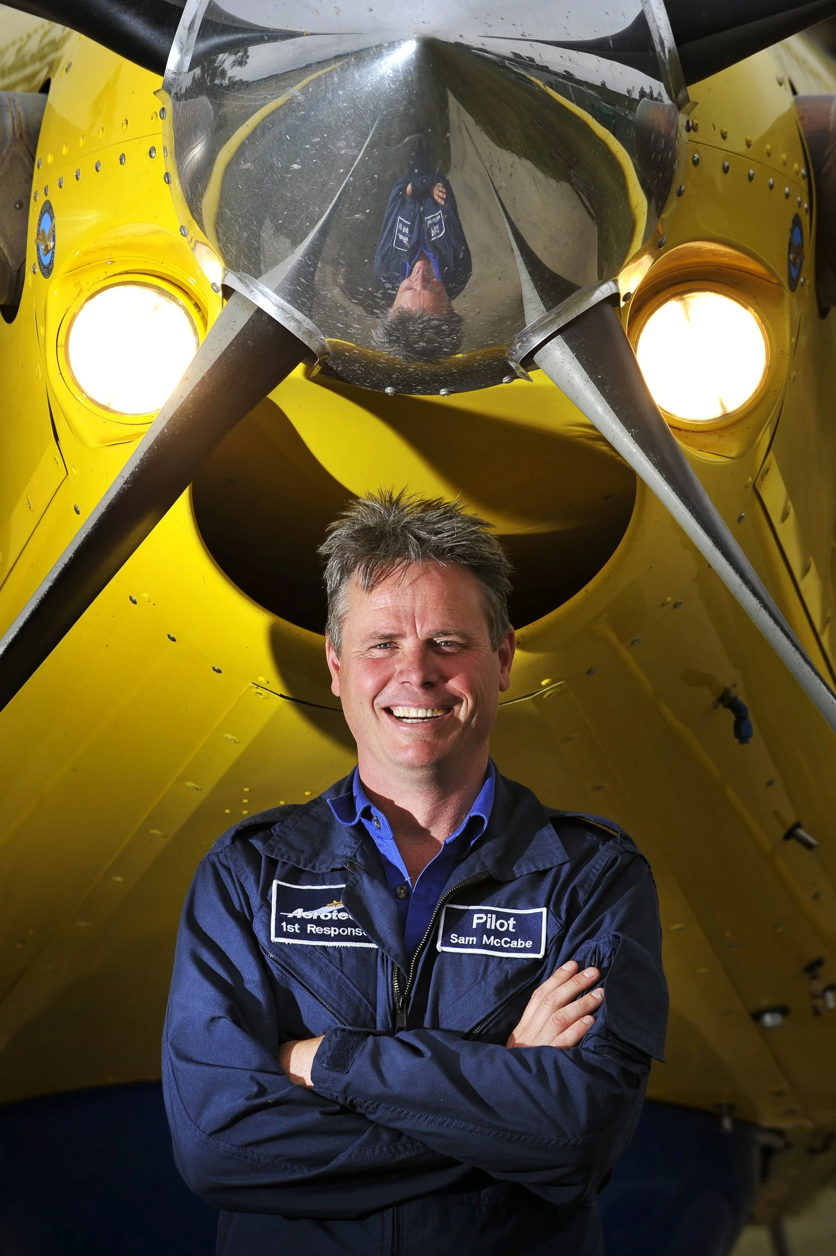 A man in a pilot uniform standing in front of a yellow aircraft, with his reflection visible in the aircraft's canopy.